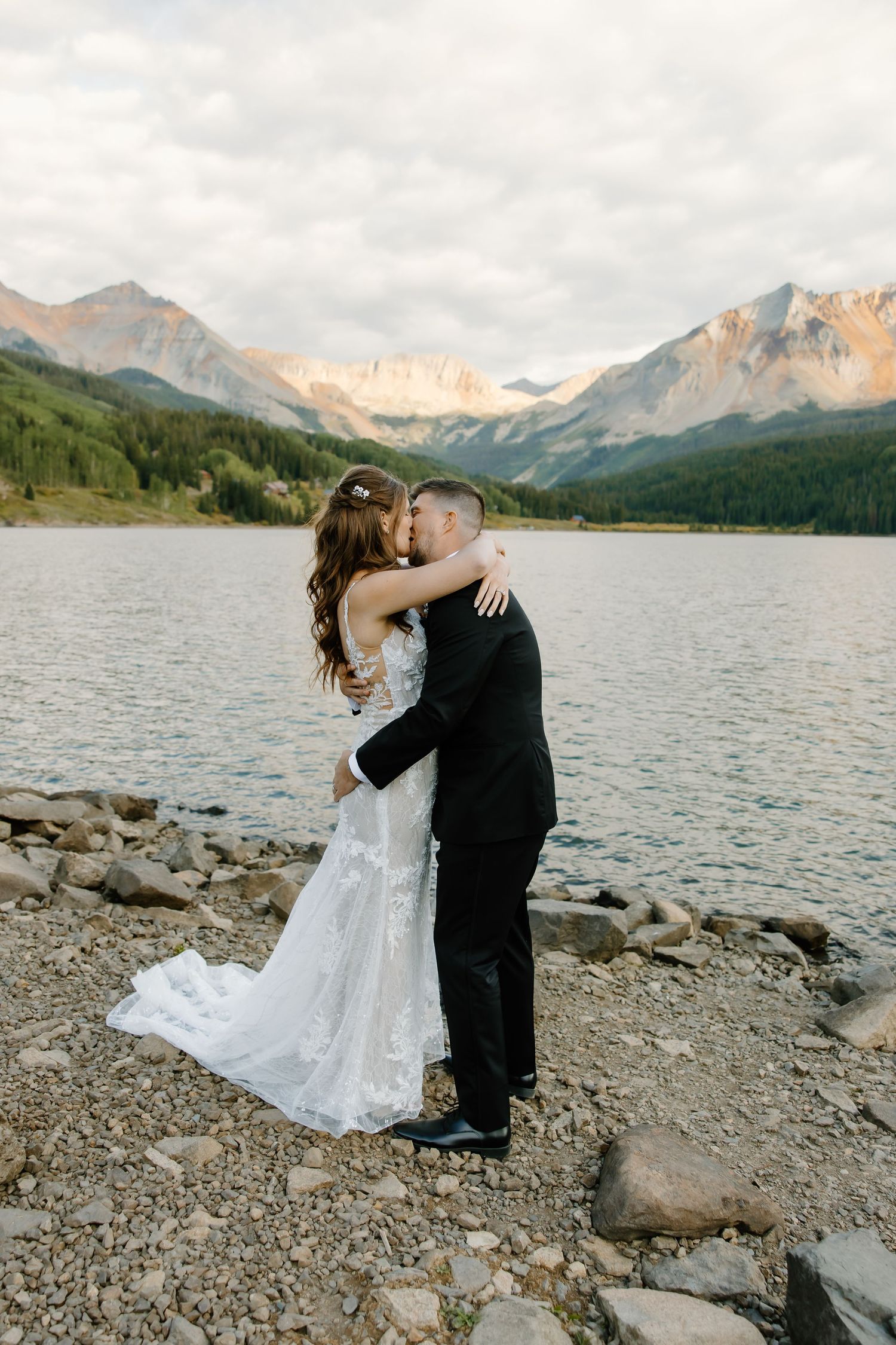 Newlyweds share an intimate moment by a mountain lake surrounded by rocky peaks.
