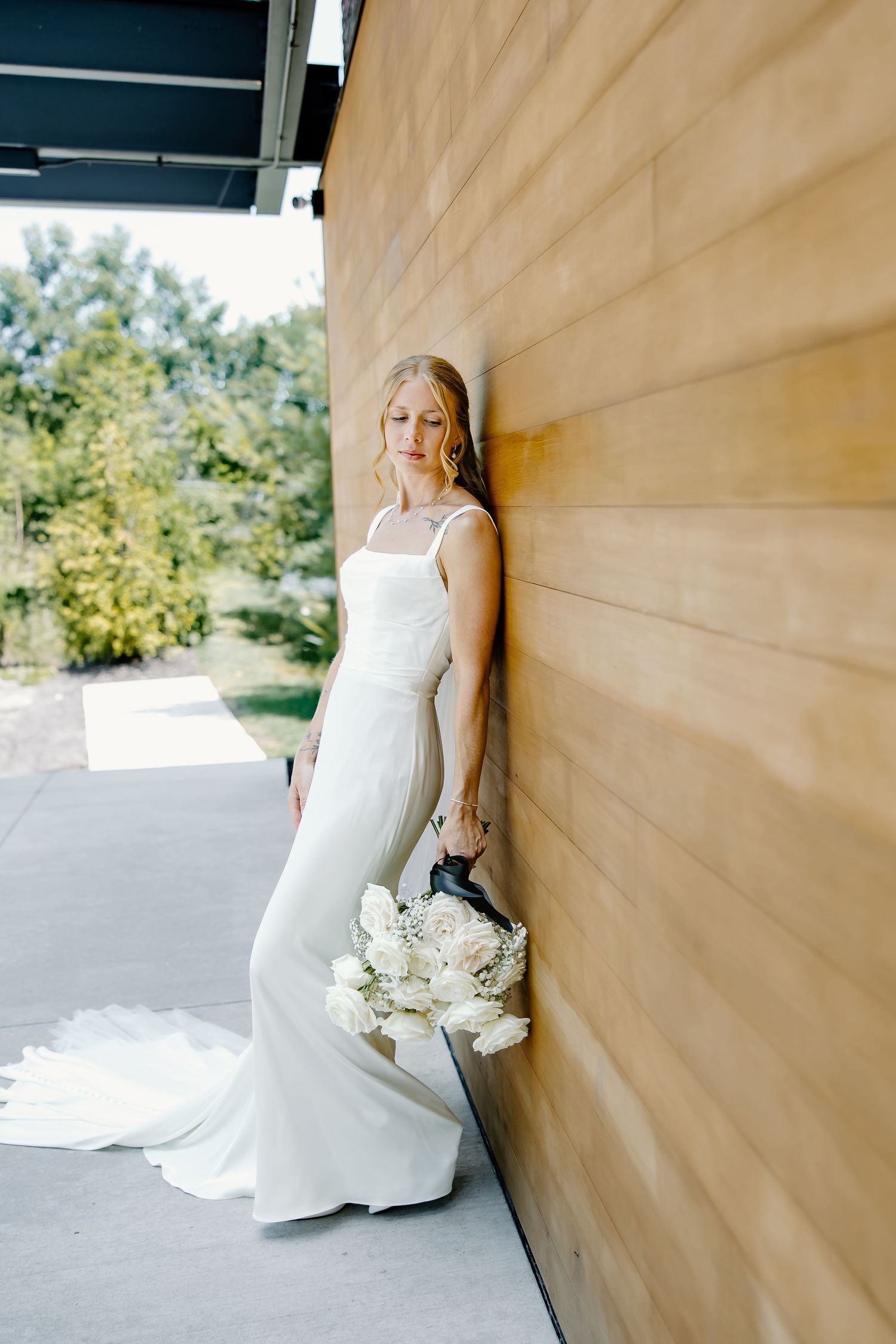 Elegant bride in sleek white gown poses against wooden wall with white bouquet.