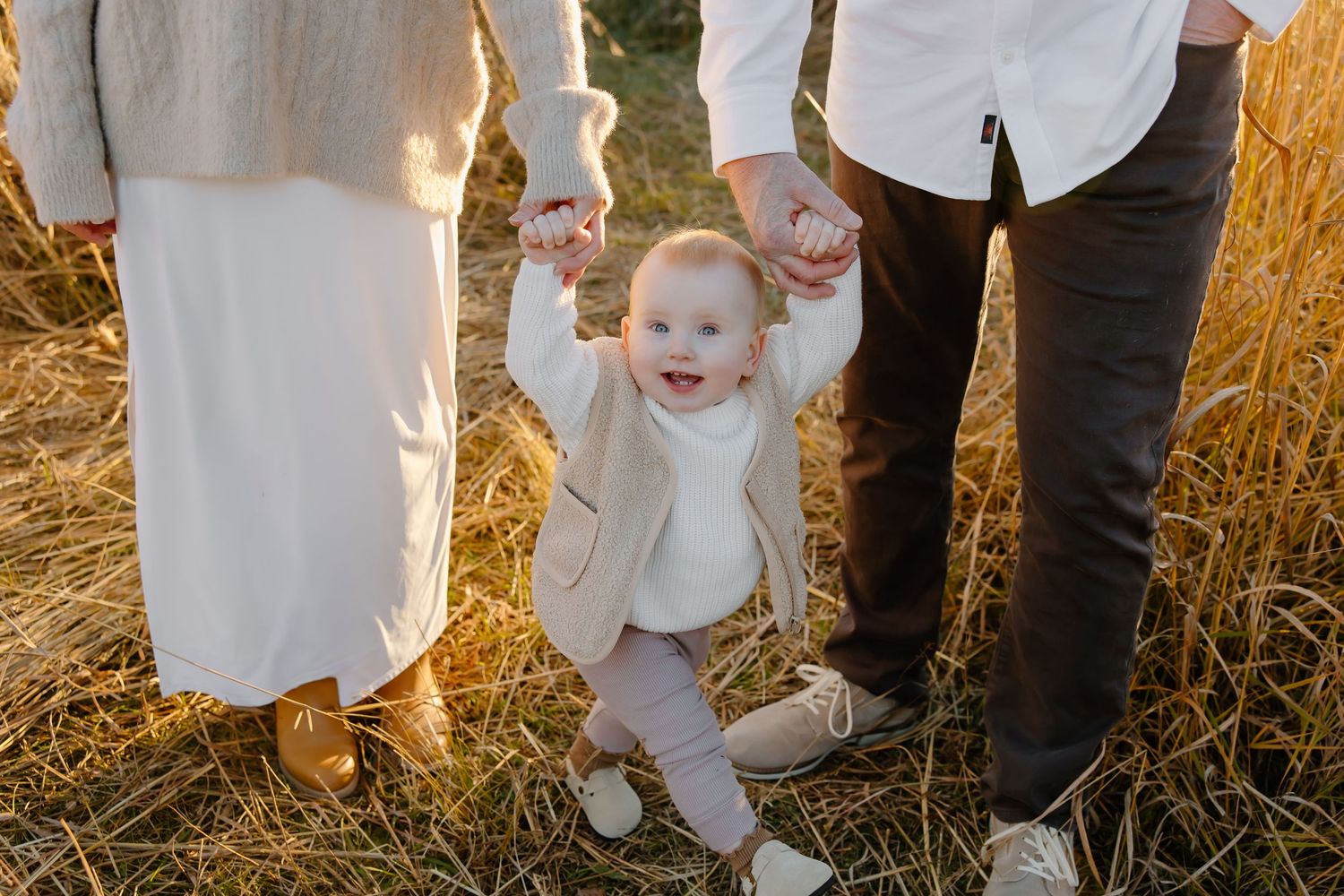 A small child in white clothing walks while holding hands with adults in a sunlit field.