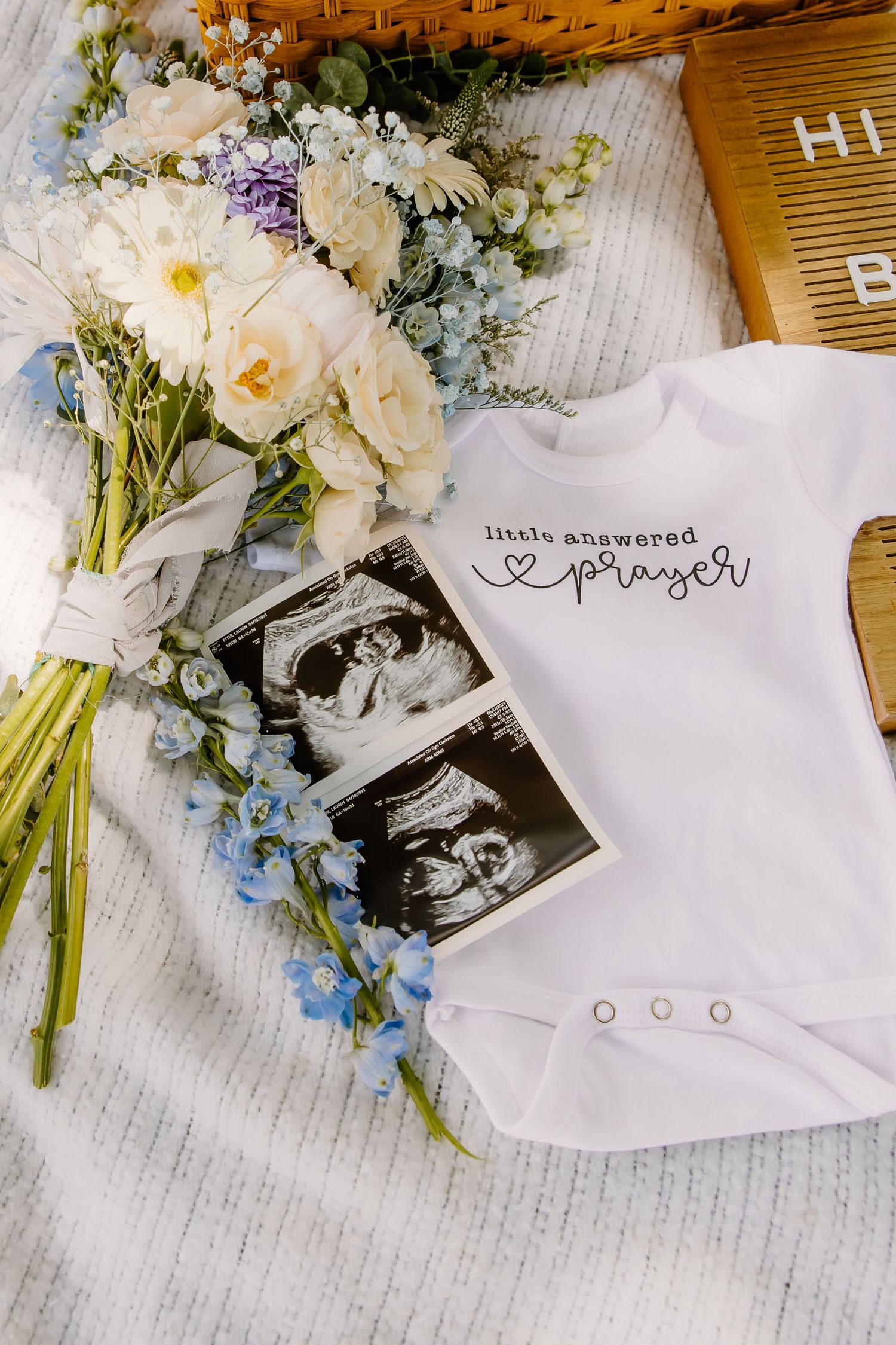 Wedding bouquet with white and blue flowers alongside a baby onesie and ultrasound photo.