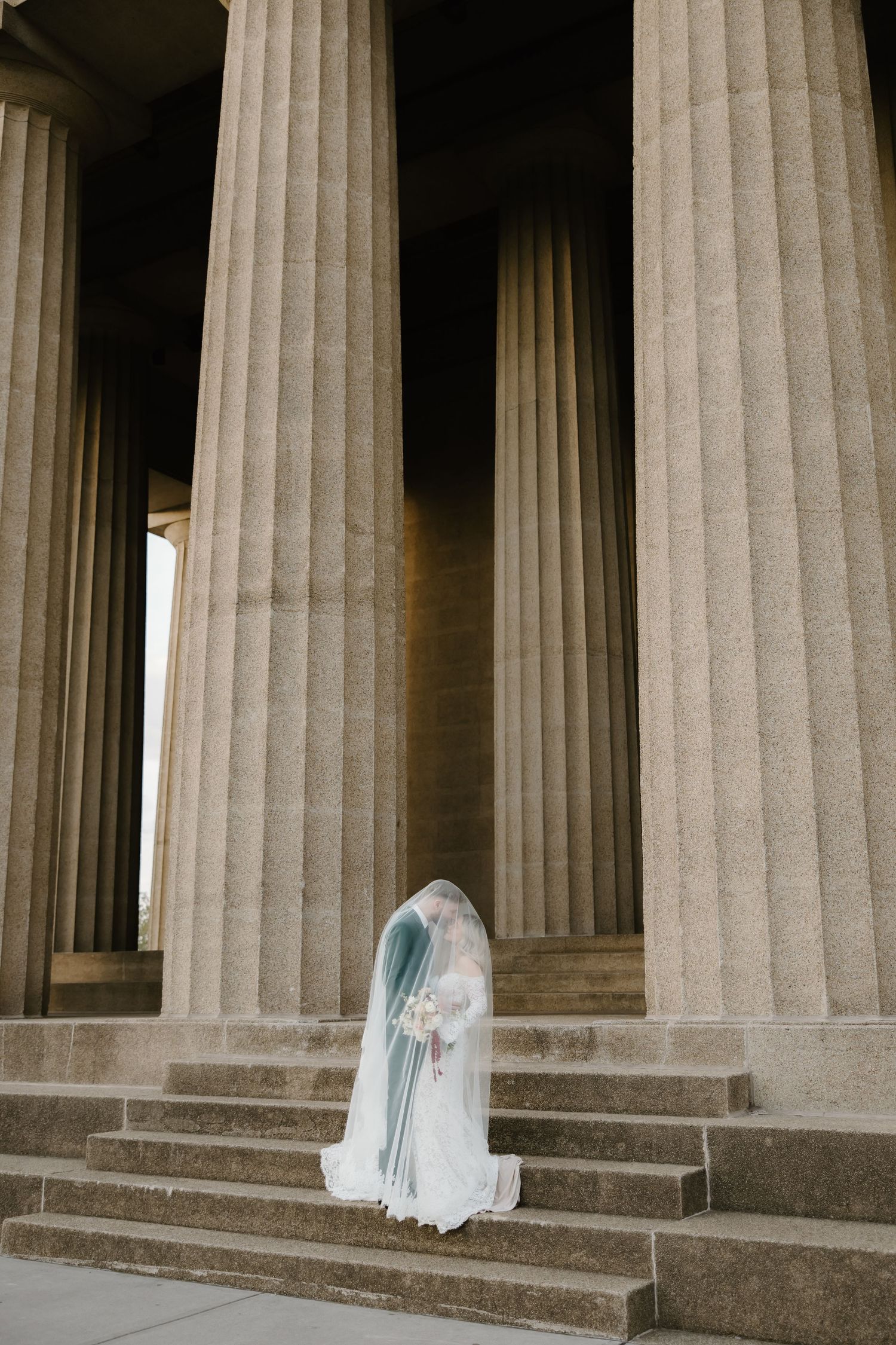Bride in white gown and veil stands on steps of neoclassical building with tall fluted columns.