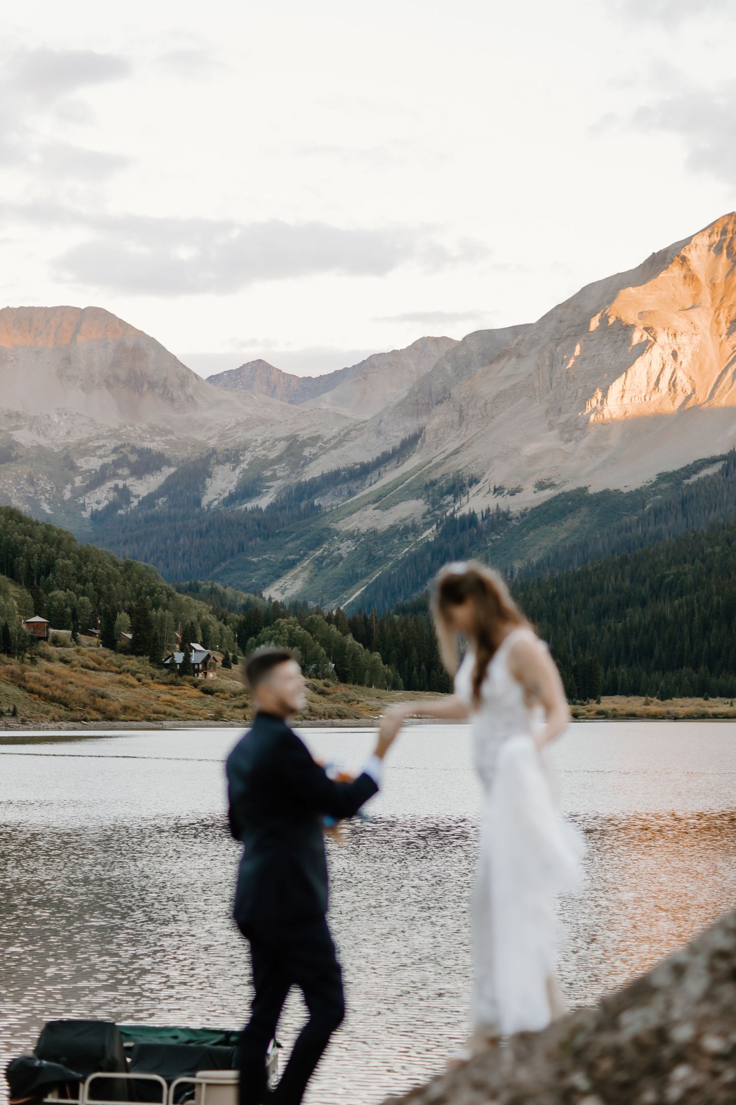 A couple in wedding attire dances playfully by a scenic mountain lake at sunset with dramatic peaks in the background.