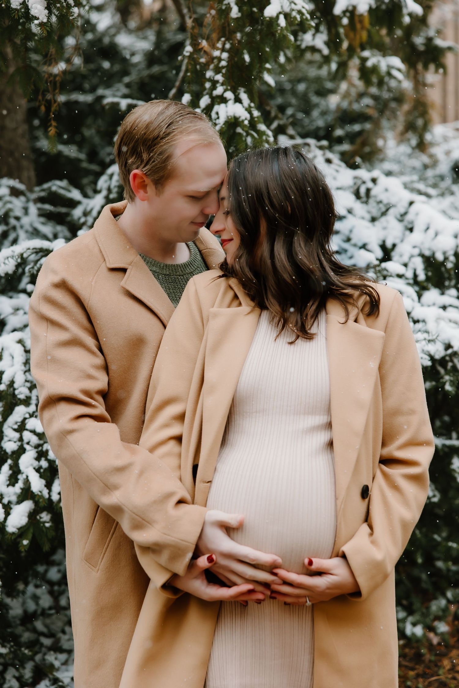 Couple in matching camel coats embrace in a snowy winter scene with the expectant mother wearing a cream dress.