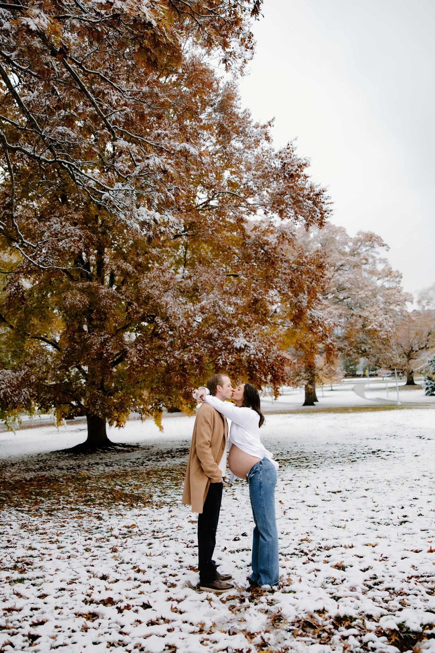 Couple walking through a snowy landscape with autumn trees in the background.