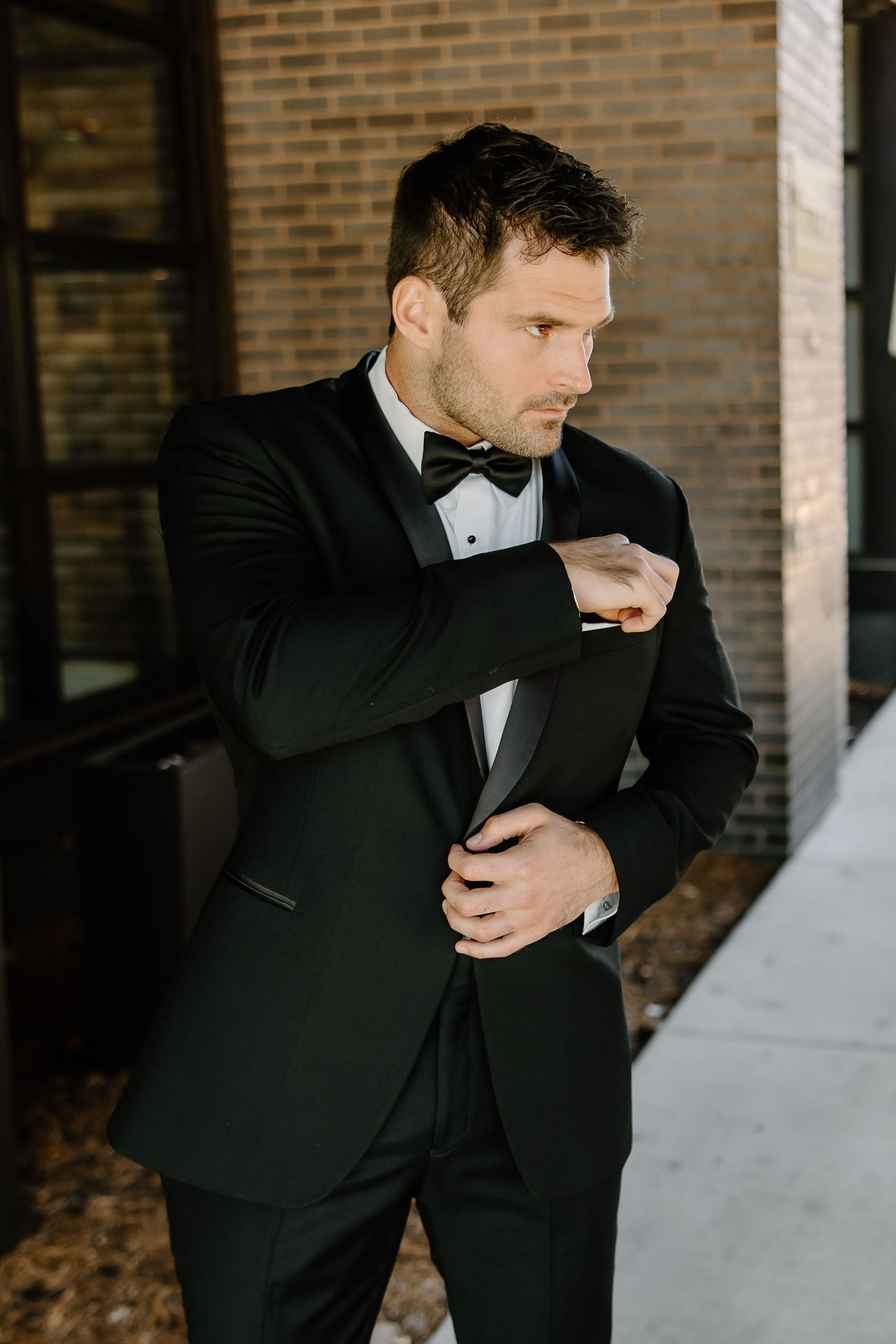 A groom in a classic black tuxedo adjusts his sleeve cuff against a brick wall backdrop.
