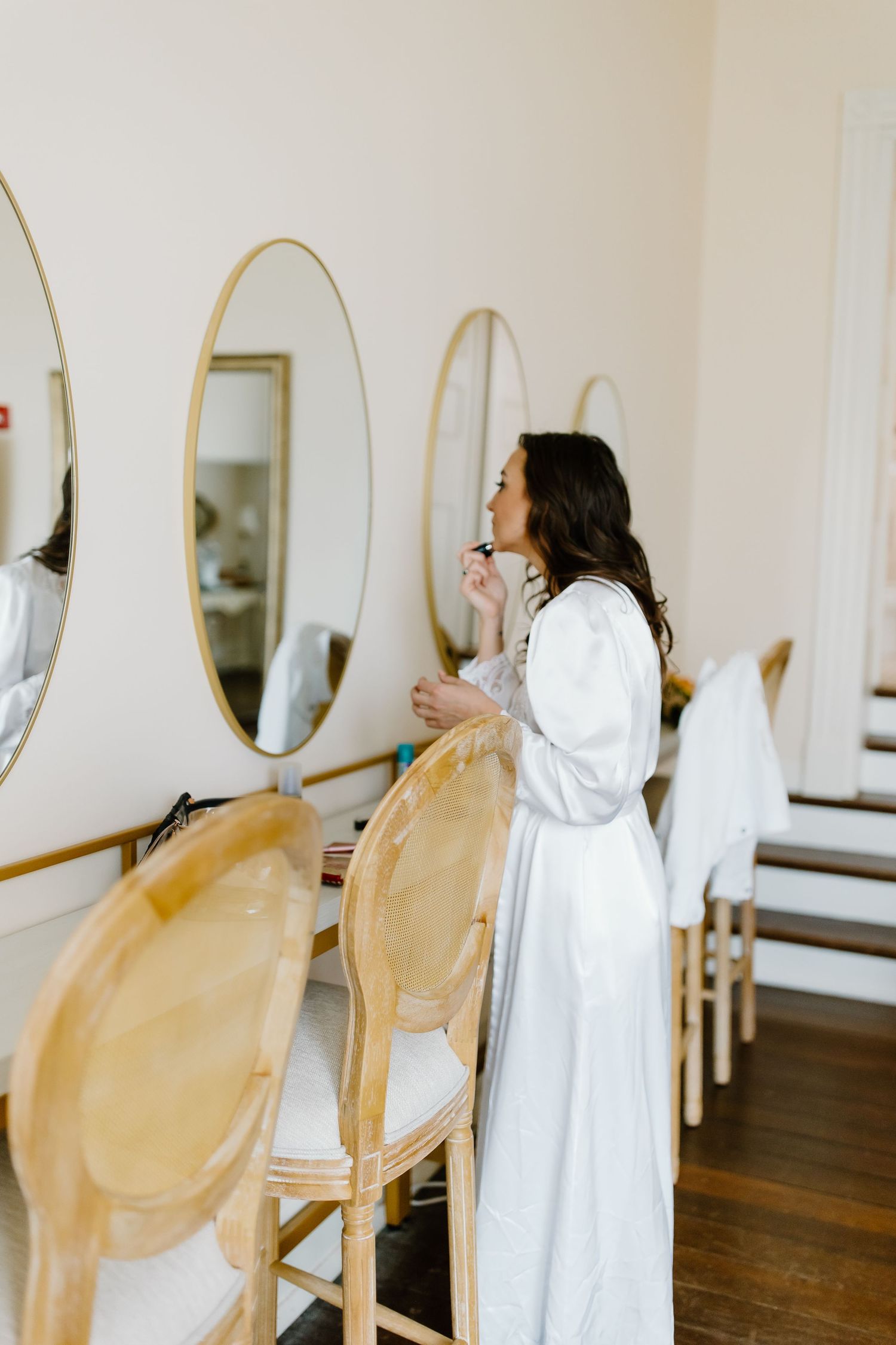 Getting ready room with gold chairs and oval mirrors mounted on white walls.