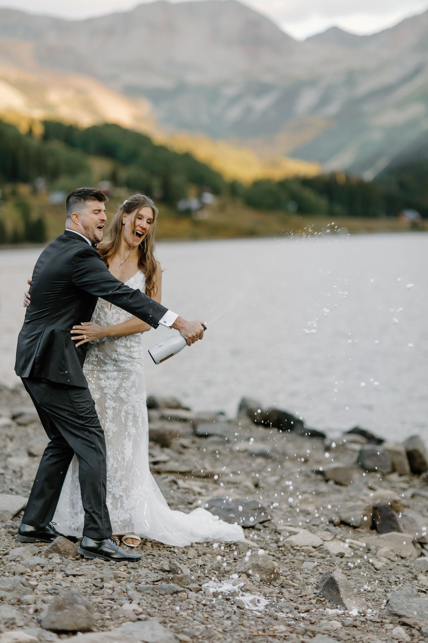 Wedding couple celebrates with champagne by a mountain lake during their photo session.