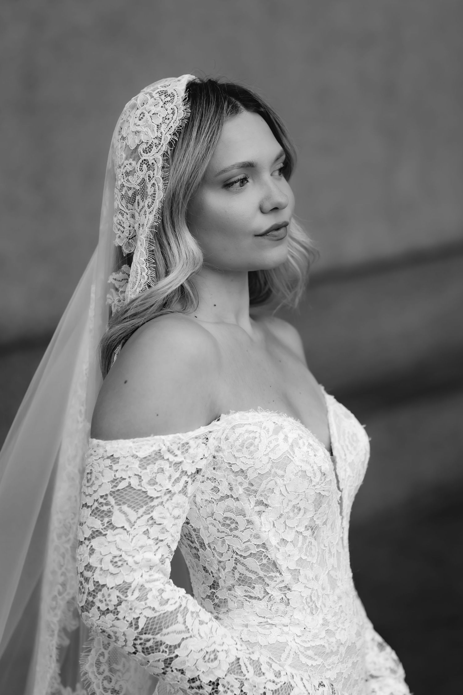 Black and white portrait of a bride in lace off-shoulder gown and lace veil, gazing elegantly to the side.