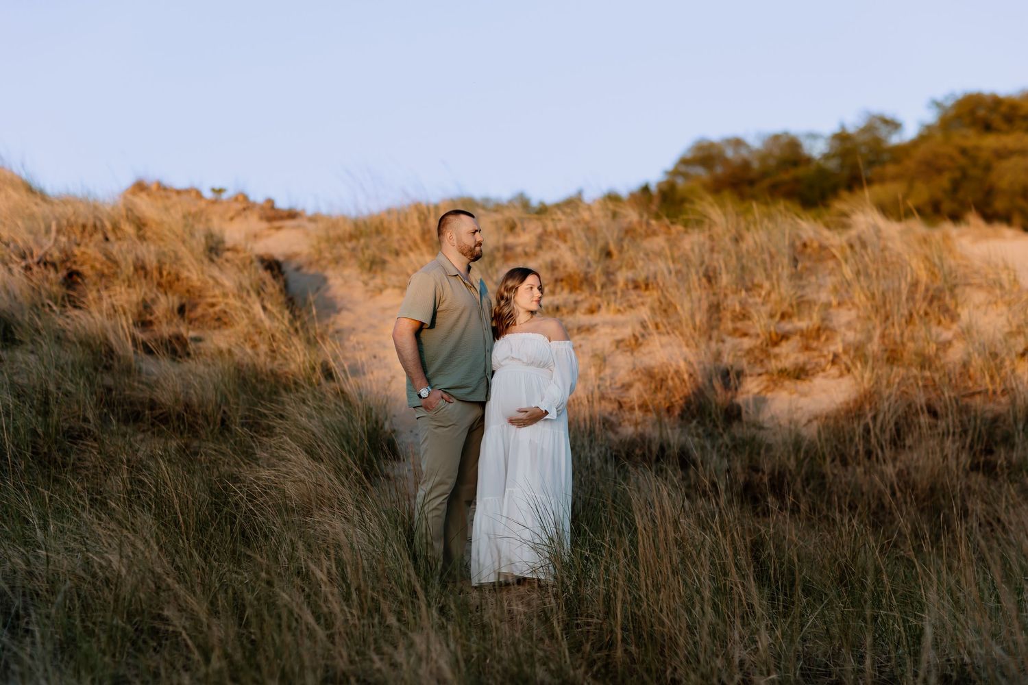 Couple standing in golden grass fields during maternity photoshoot at sunset, woman in white dress cradling baby bump.