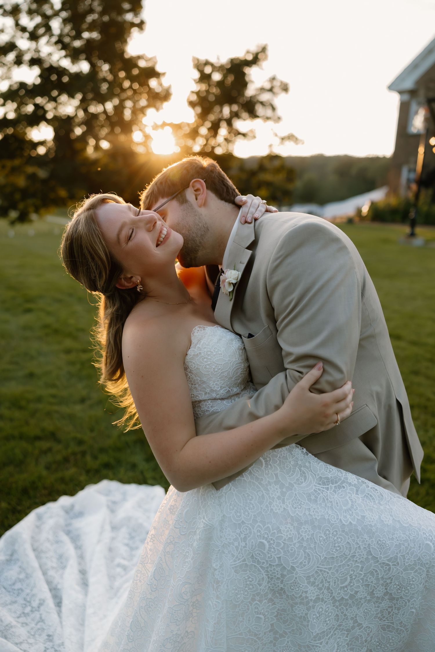 Bride and groom share a romantic sunset embrace outdoors, with golden light glowing behind them on lush green grounds.