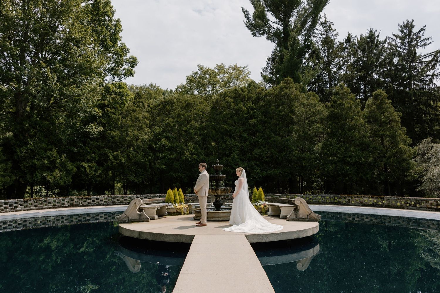 Bride and groom stand on a fountain island surrounded by a reflective pool in a lush garden wedding venue.