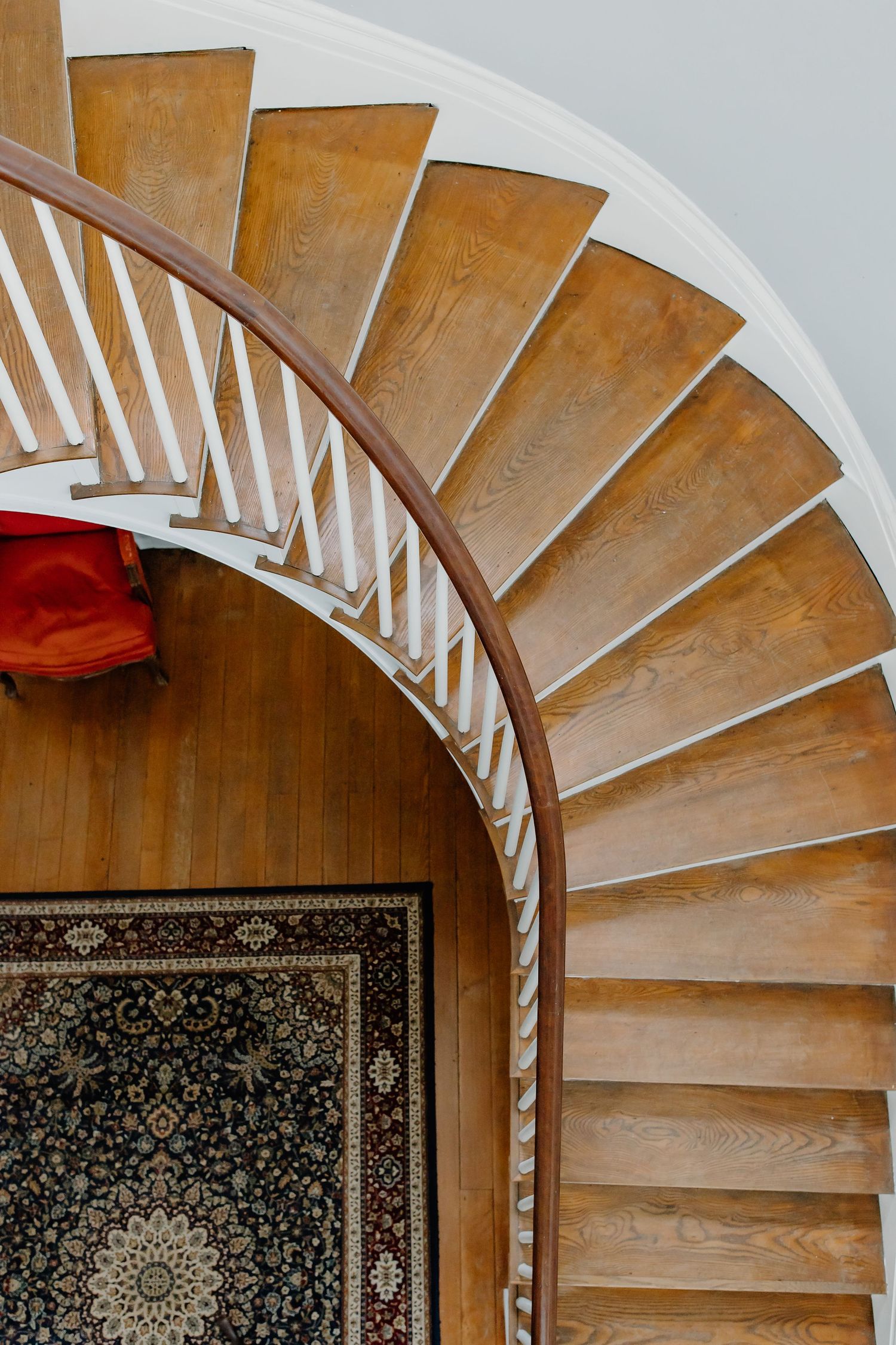 Curved wooden staircase with white spindles and ornate patterned rug at the bottom against a light wall.