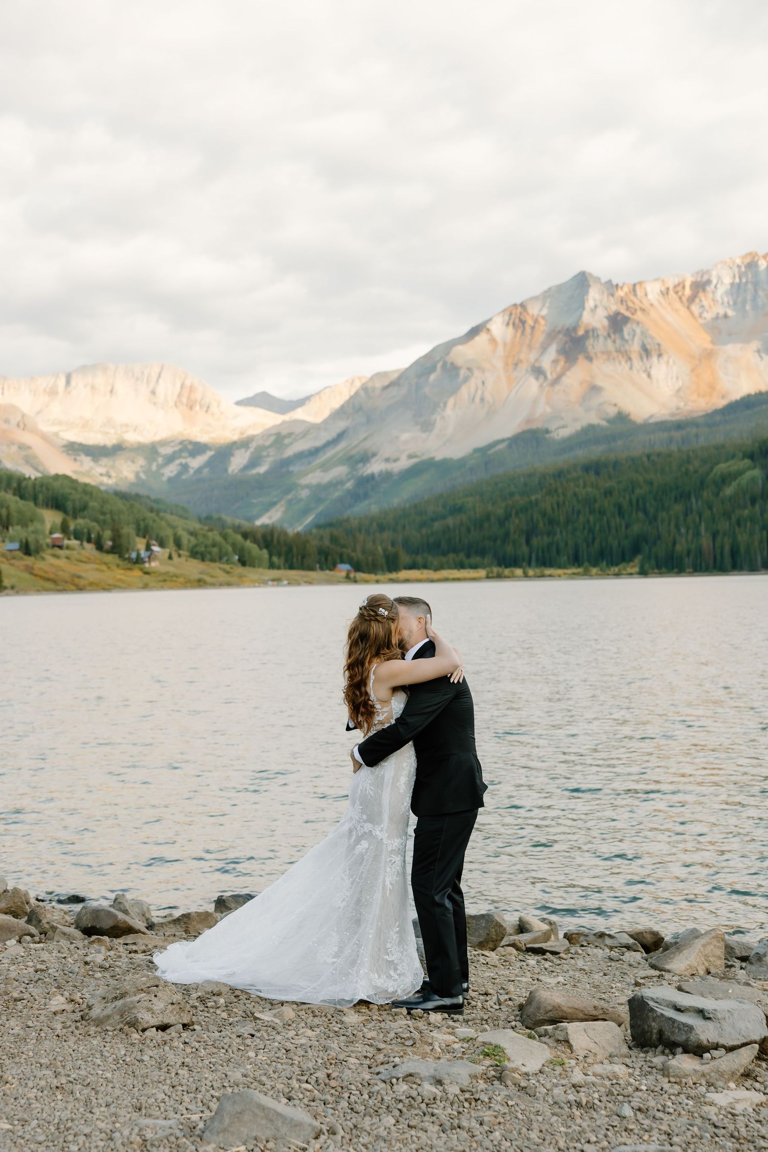 A couple embraces during their mountain wedding photoshoot beside a scenic alpine lake at sunset.