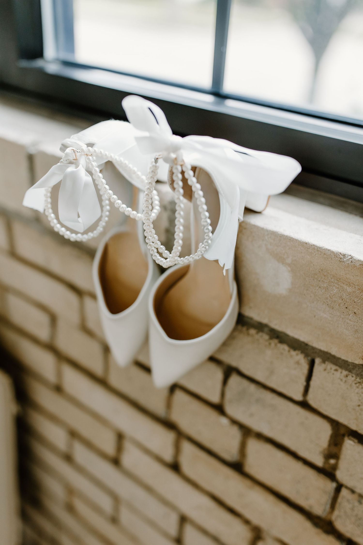 White bridal shoes with pearl details displayed on a brick windowsill.