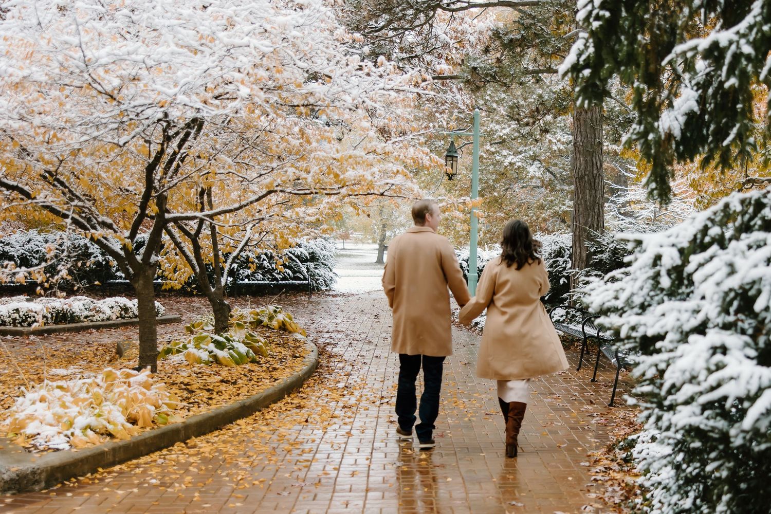 Couple walking together on brick pathway surrounded by snow and autumn leaves.