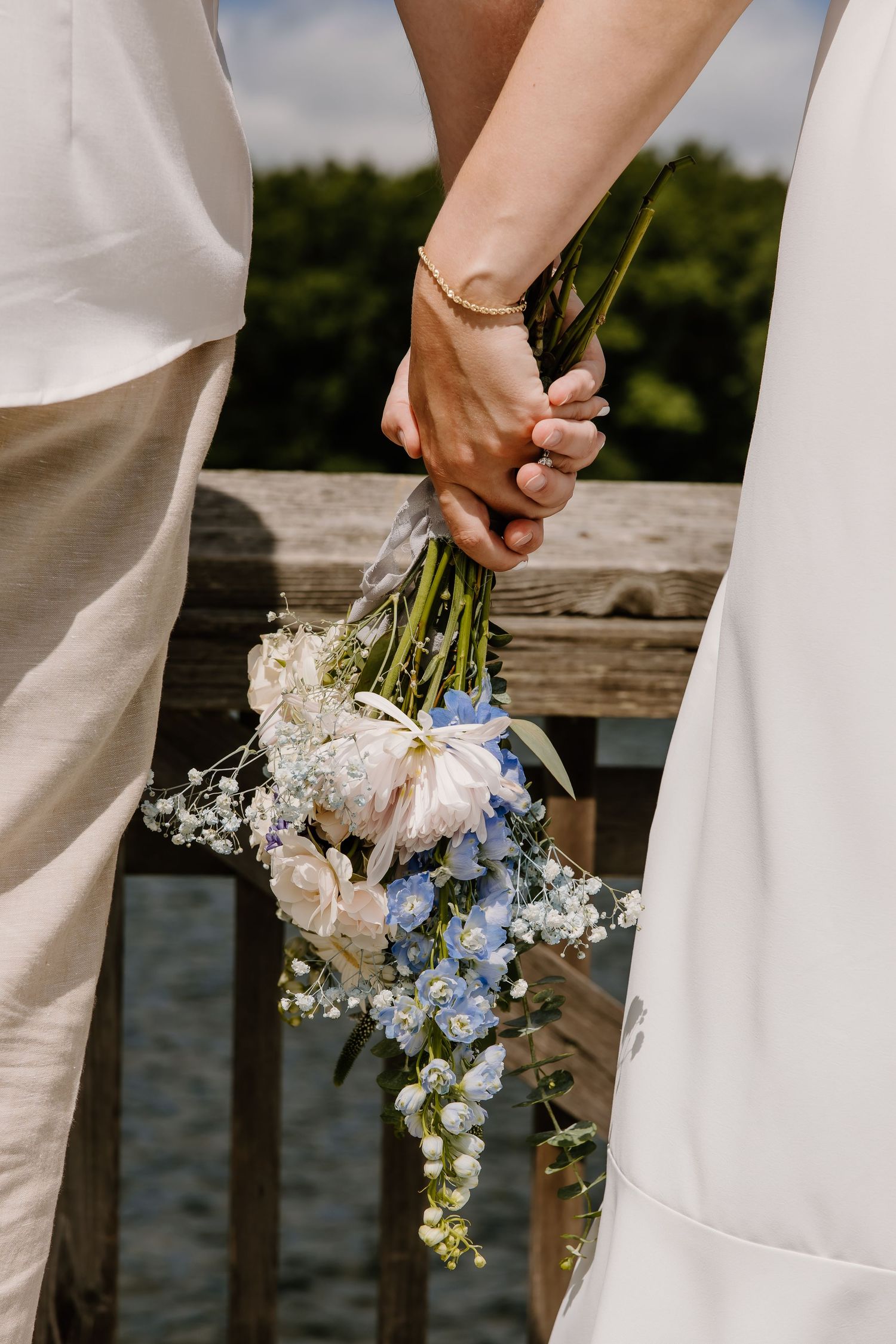Close-up of joined hands holding a delicate bouquet of blue and white flowers against wedding attire.