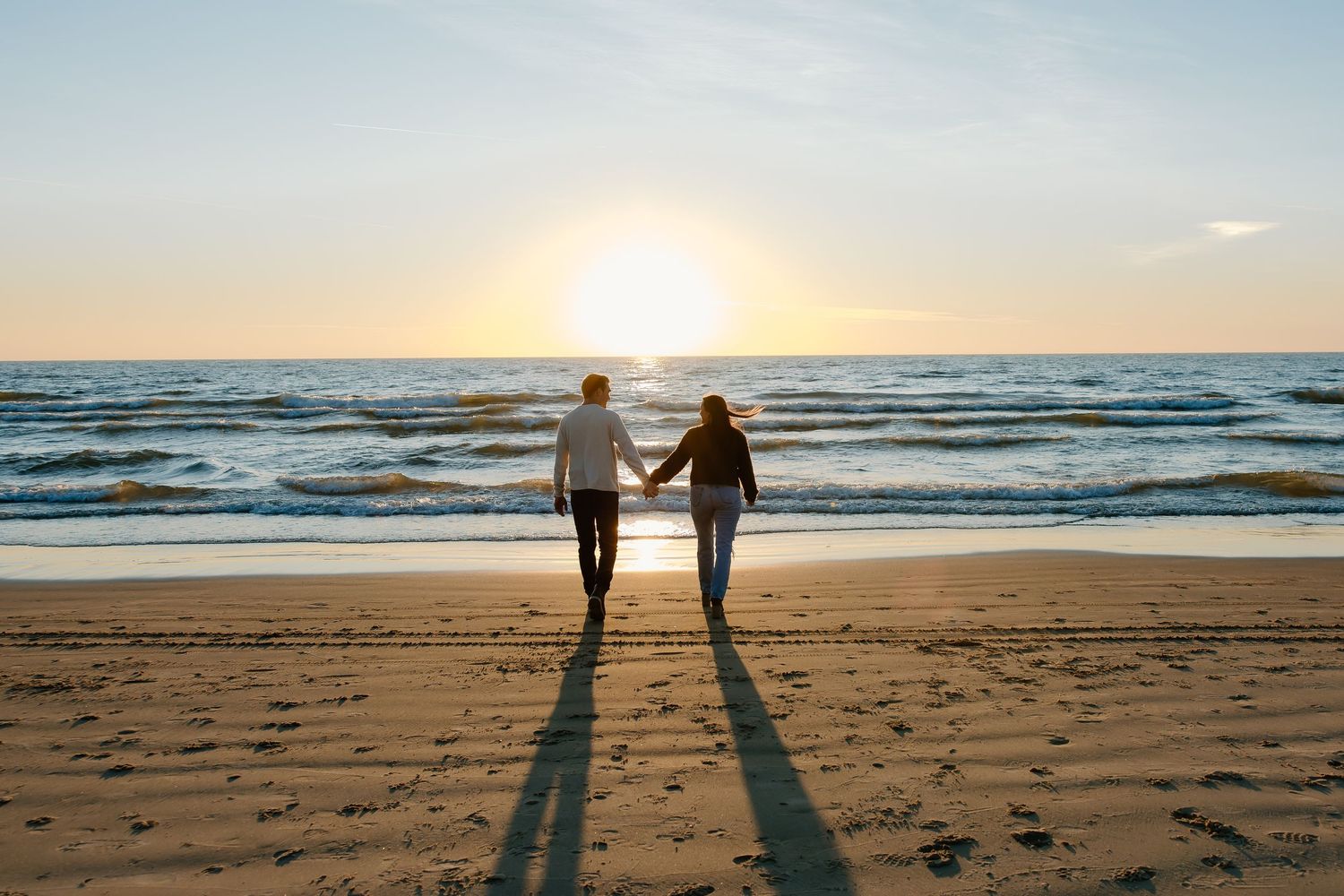 Two people standing together on a sandy beach with ocean waves and sunset in the background.