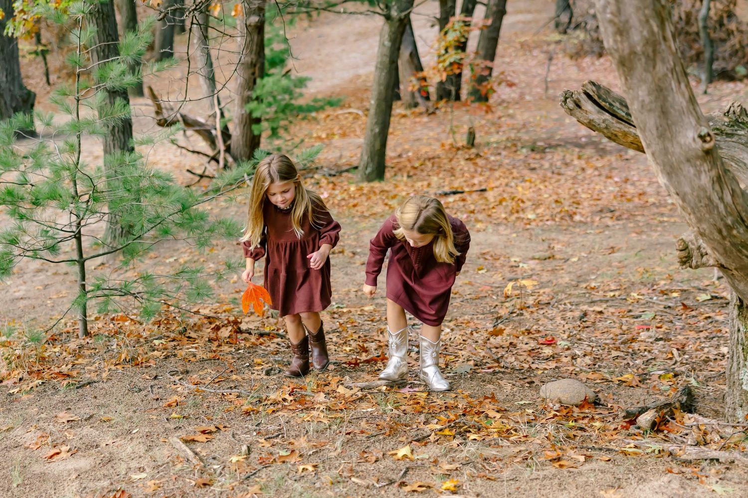 Children in matching burgundy dresses play together on sandy ground with fall foliage in background.