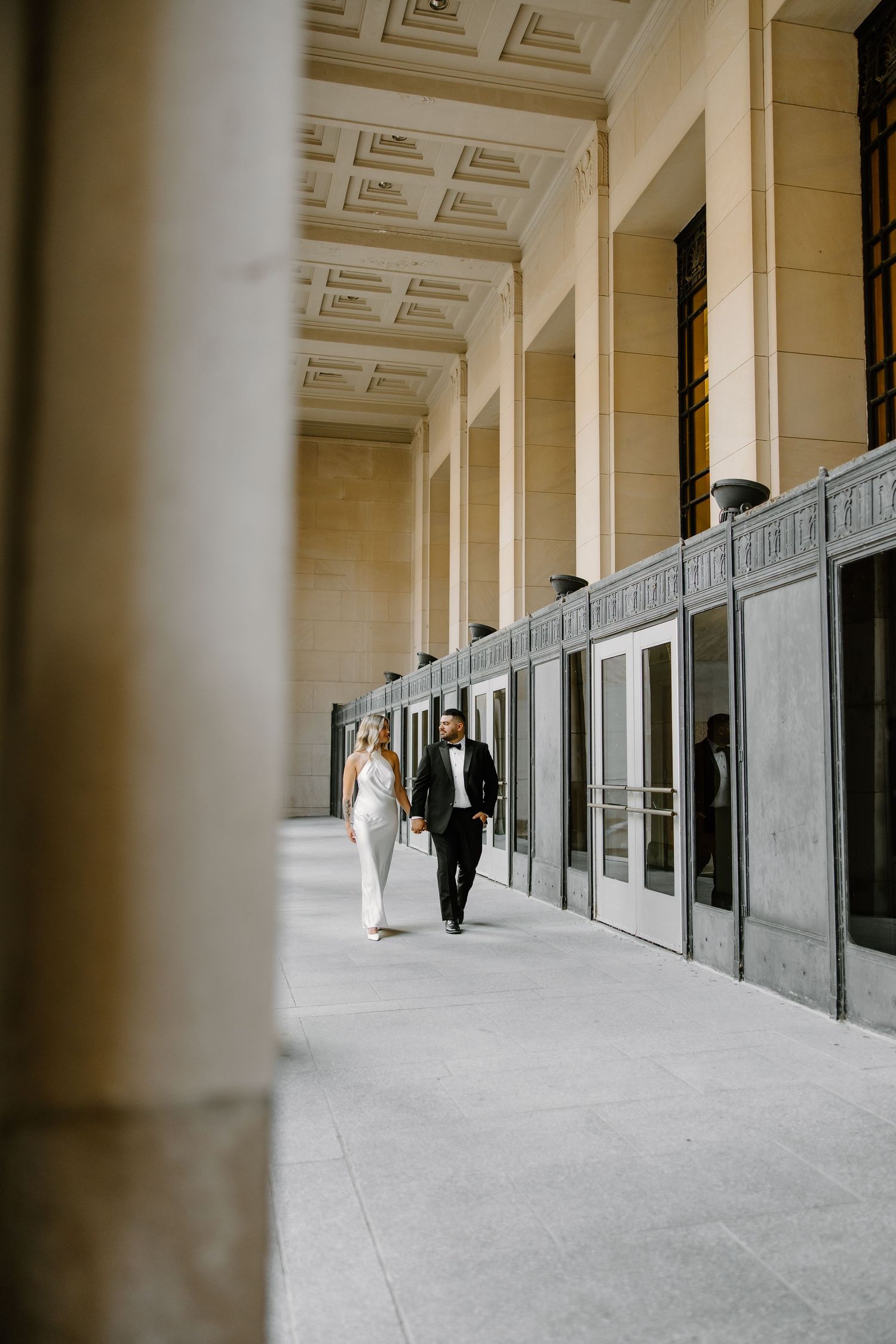 Couple in formal wear walks through classical architectural corridor.