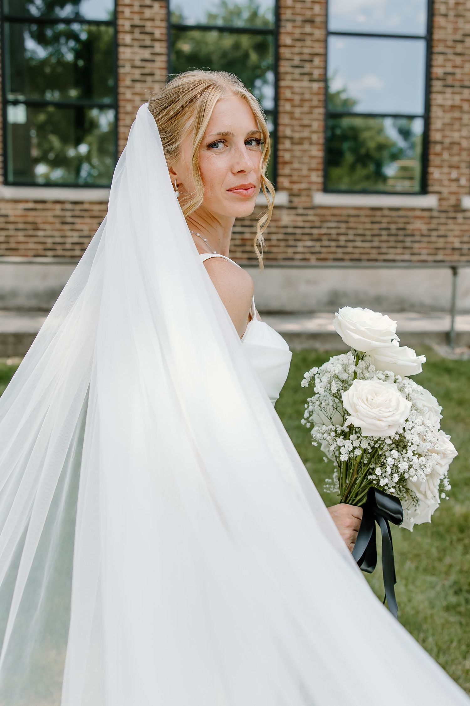 Elegant bridal portrait with white roses and flowing veil against a brick building backdrop.