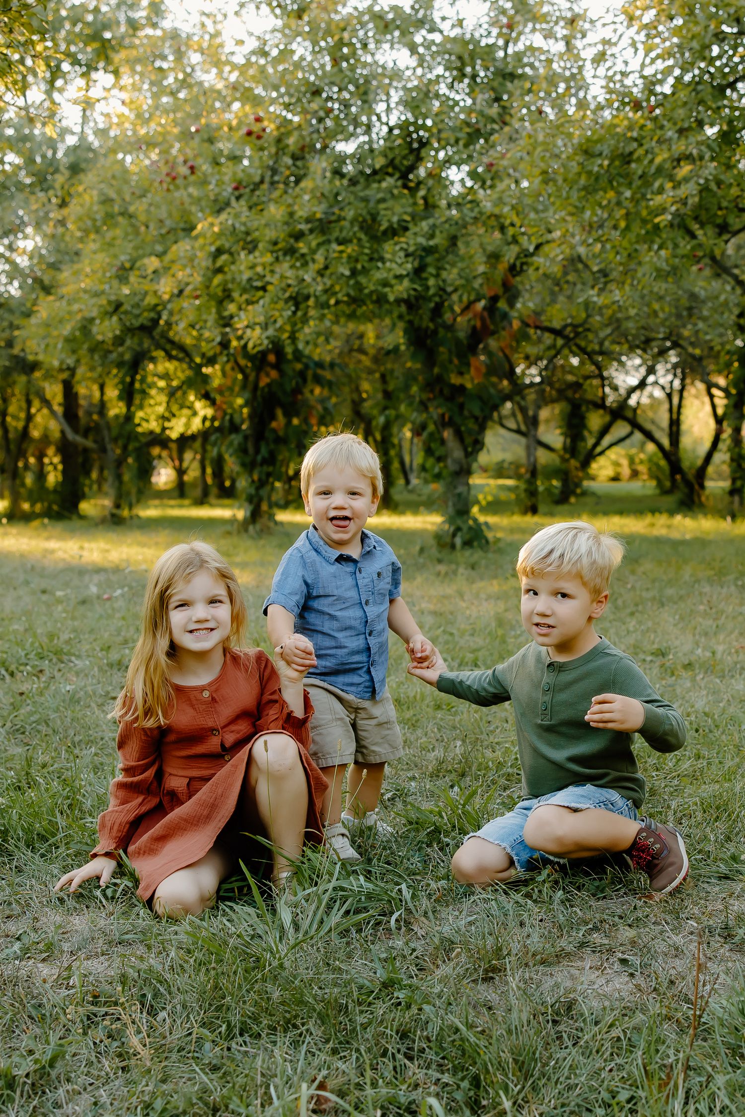 Three children sitting together on grass in an orchard during golden hour.