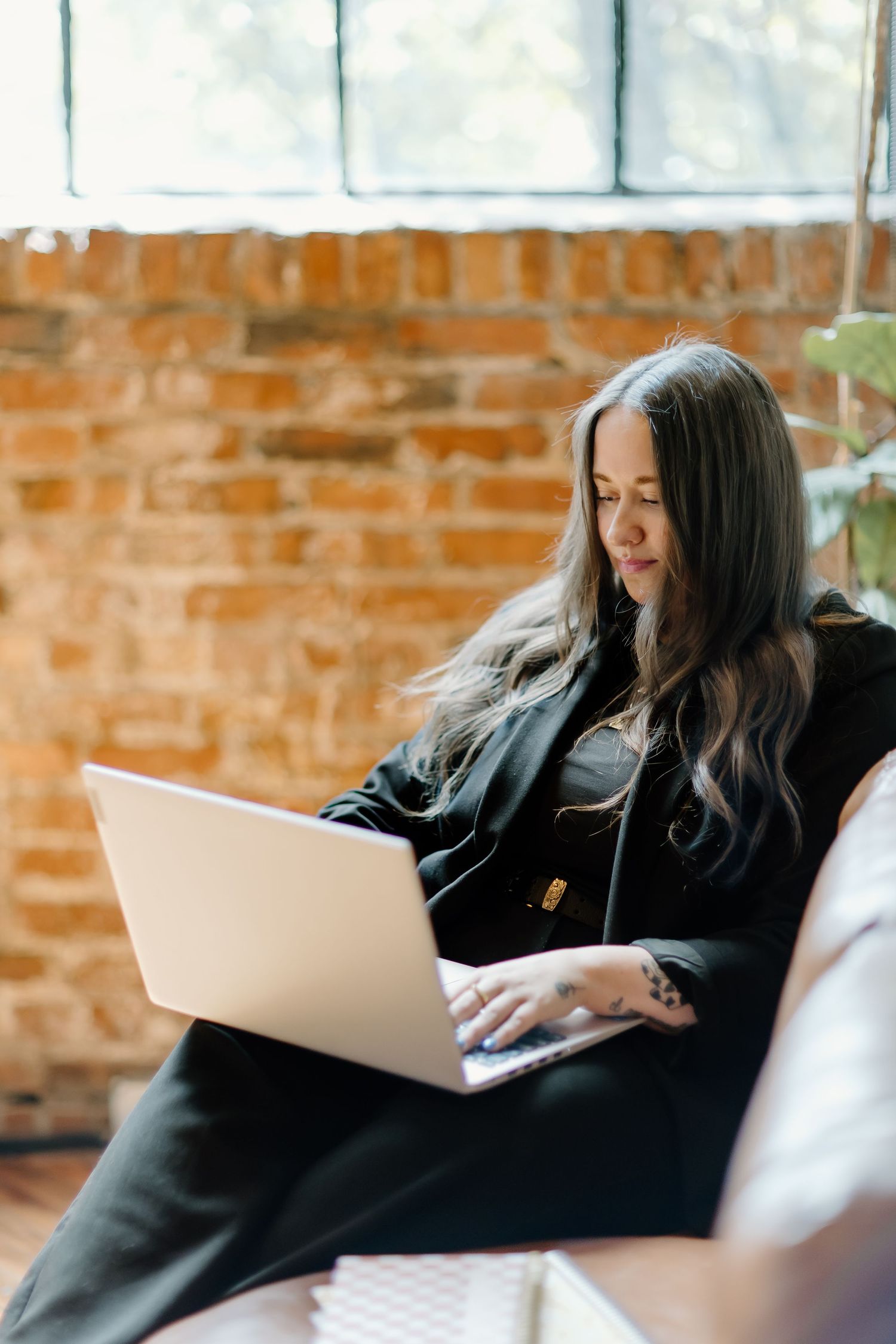 Person in black sweater working on laptop in brick-walled room.