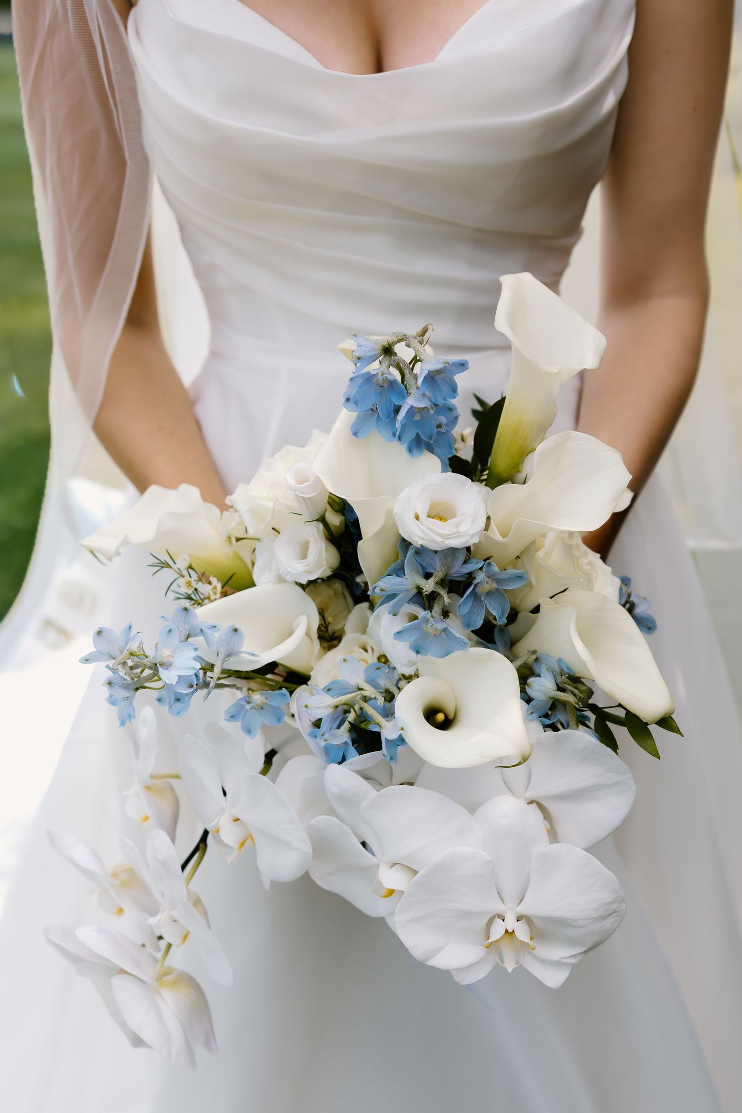 Bride in white gown holding elegant wedding bouquet of white calla lilies, orchids, and blue delphinium flowers.