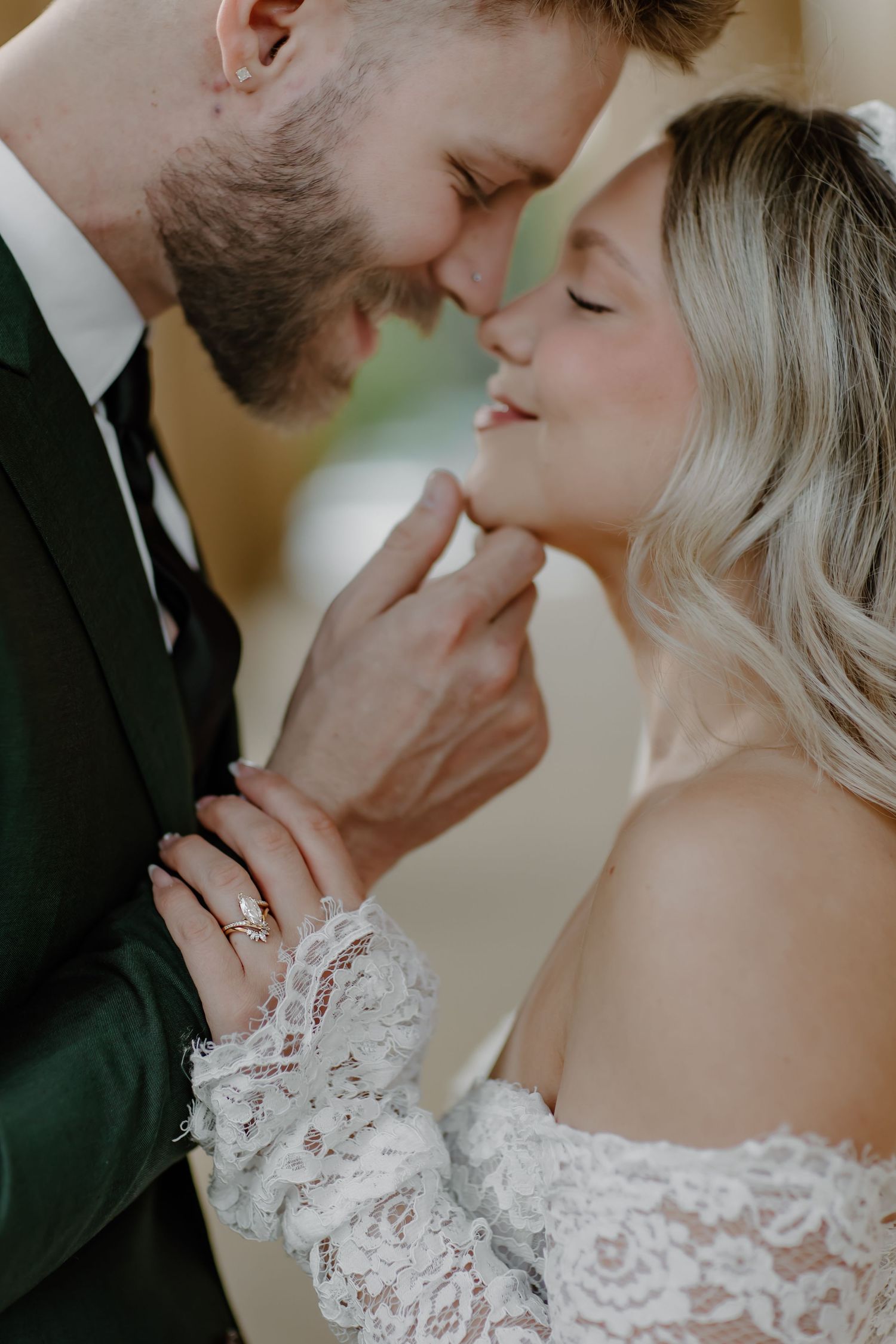 A bride and groom share a tender moment, nearly kissing, as she wears a lace off-shoulder gown and diamond ring.