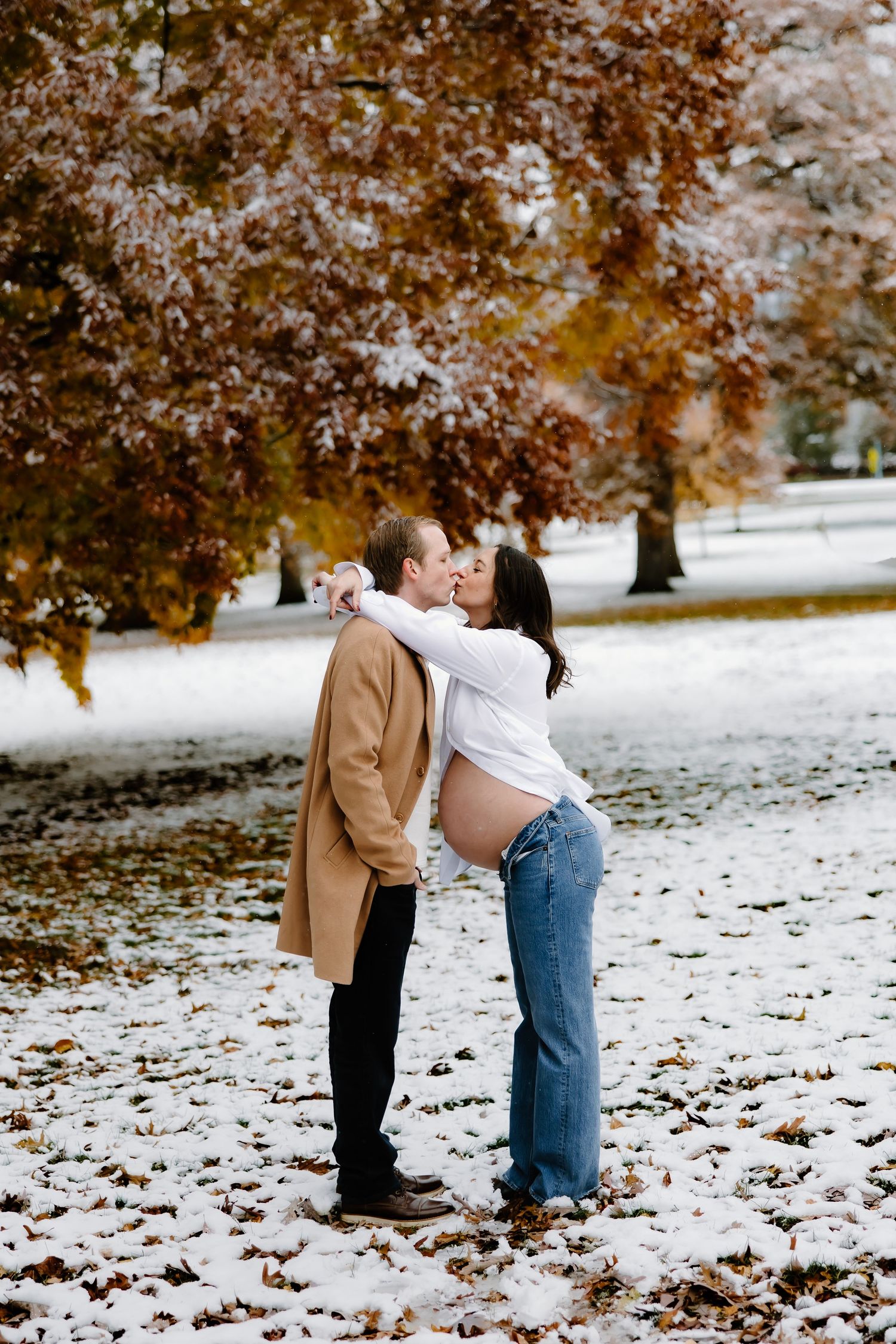 A couple embraces in a snowy park with autumn leaves and trees in the background.