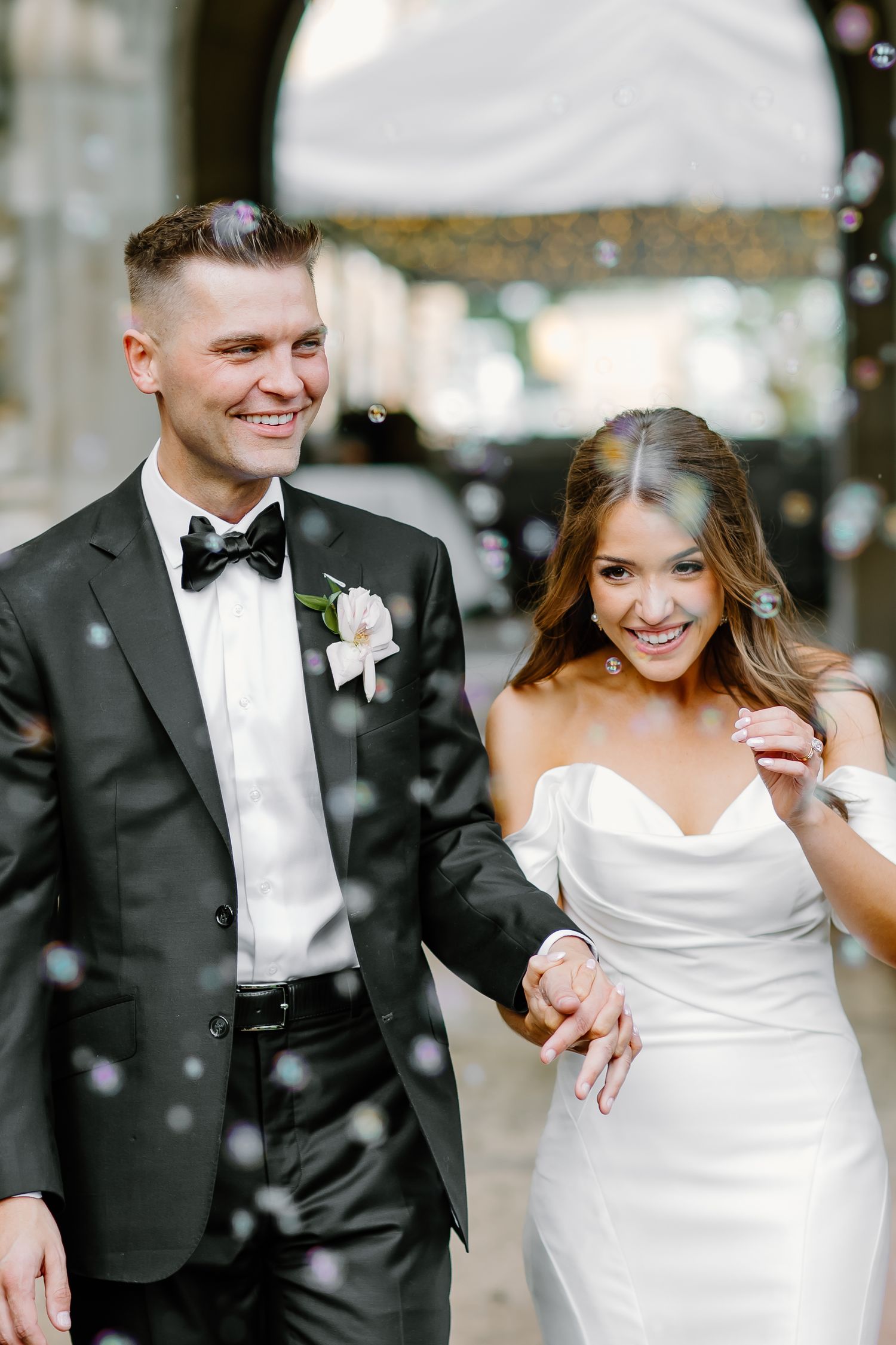 A joyful wedding couple laughs together while confetti falls around them in an elegant indoor setting.