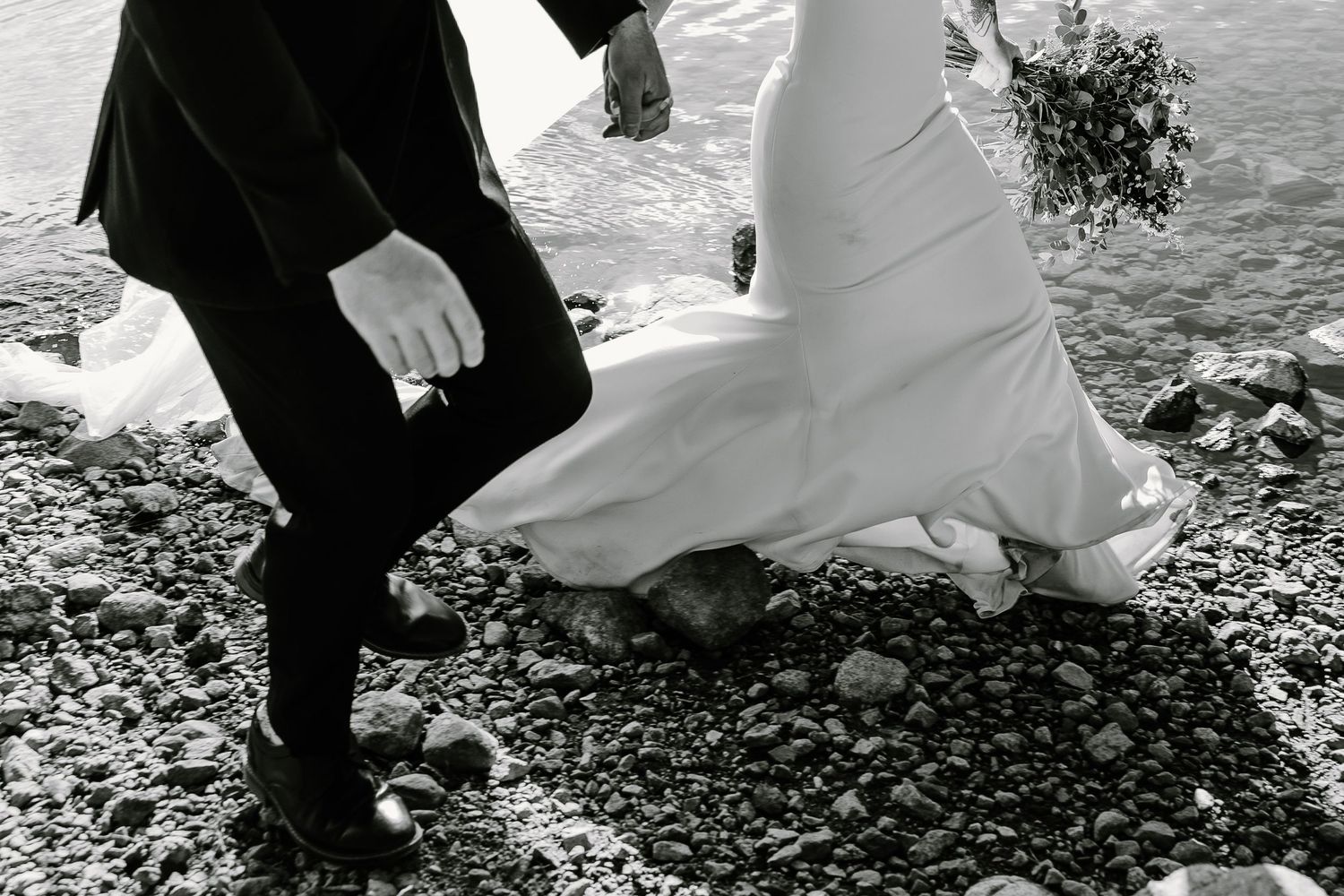 Black and white photo of couple walking on rocky beach during wedding photoshoot.