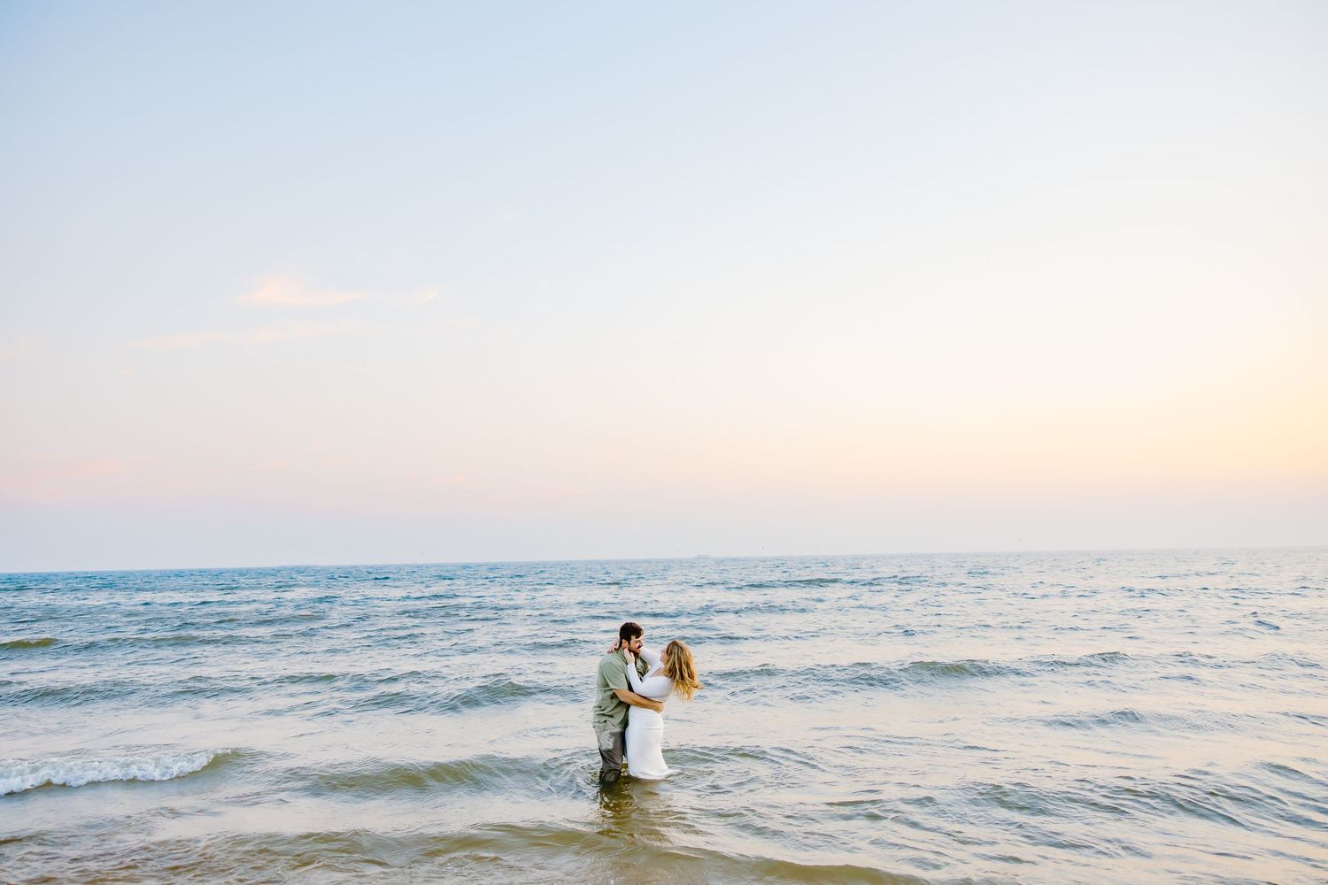 A couple stands together by the ocean during golden hour, creating silhouettes against the setting sun.