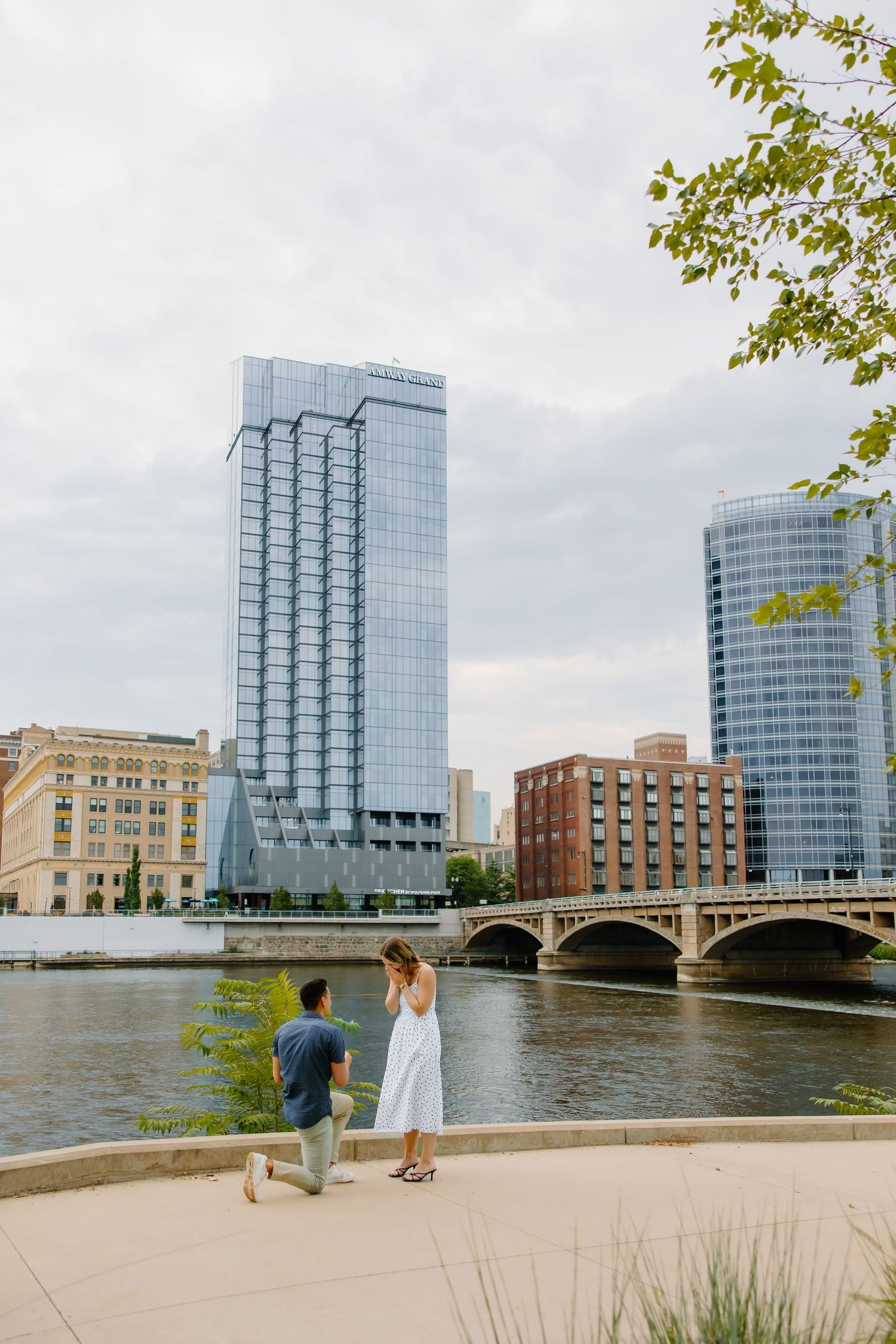 Scenic waterfront proposal scene with modern glass buildings and historic bridge in background.