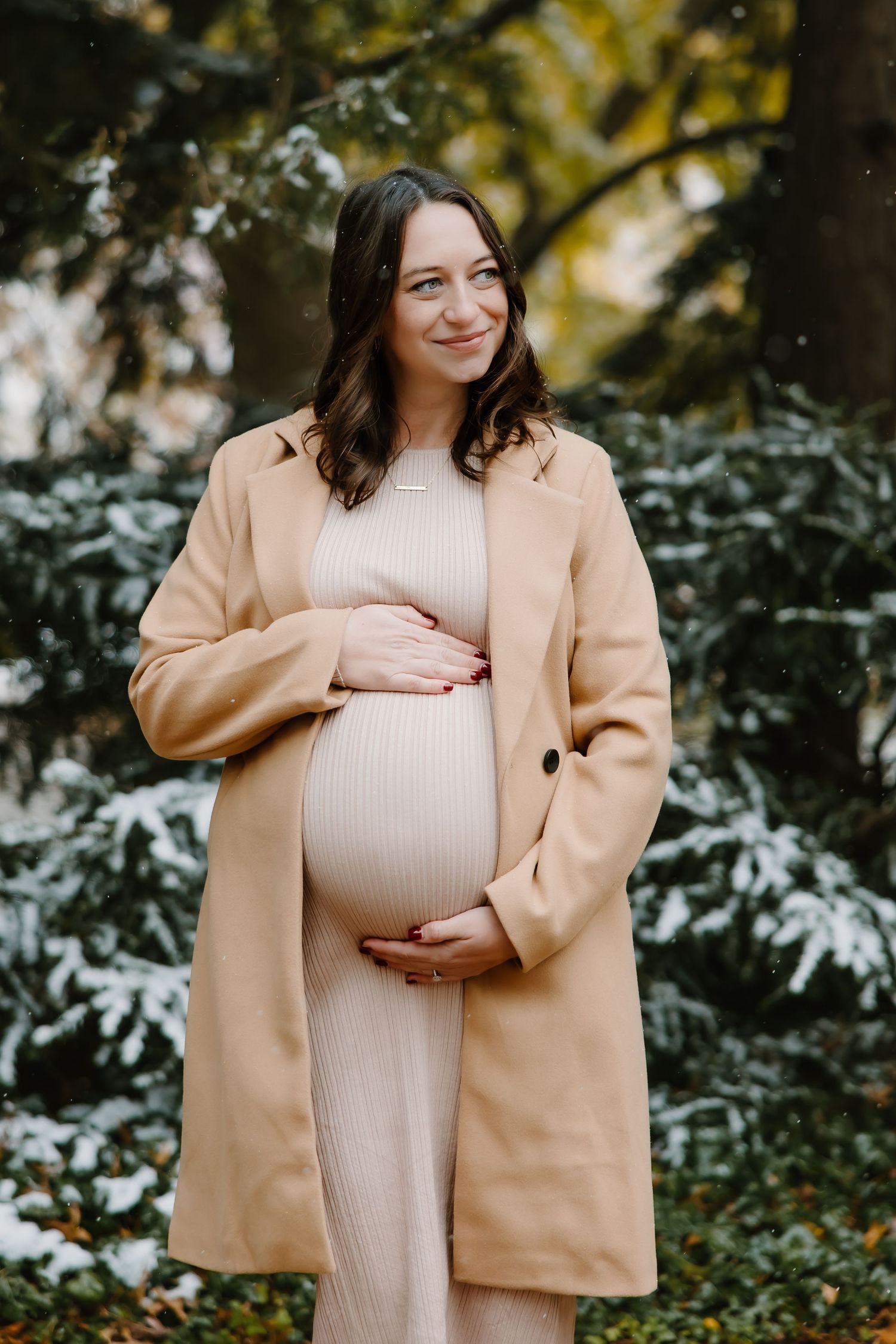 Maternity photos showing an expectant mother in a cream dress and camel coat against snowy scenery.