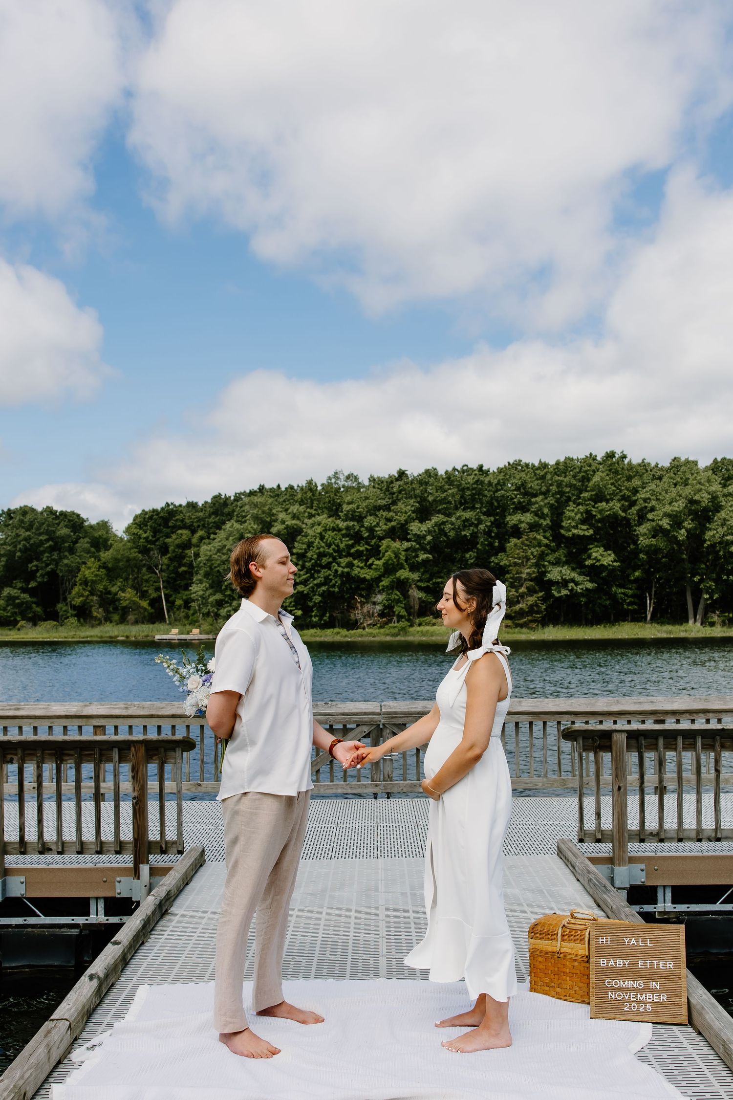 A couple stands together on a dock by a lake, dressed in white for an intimate wedding ceremony.