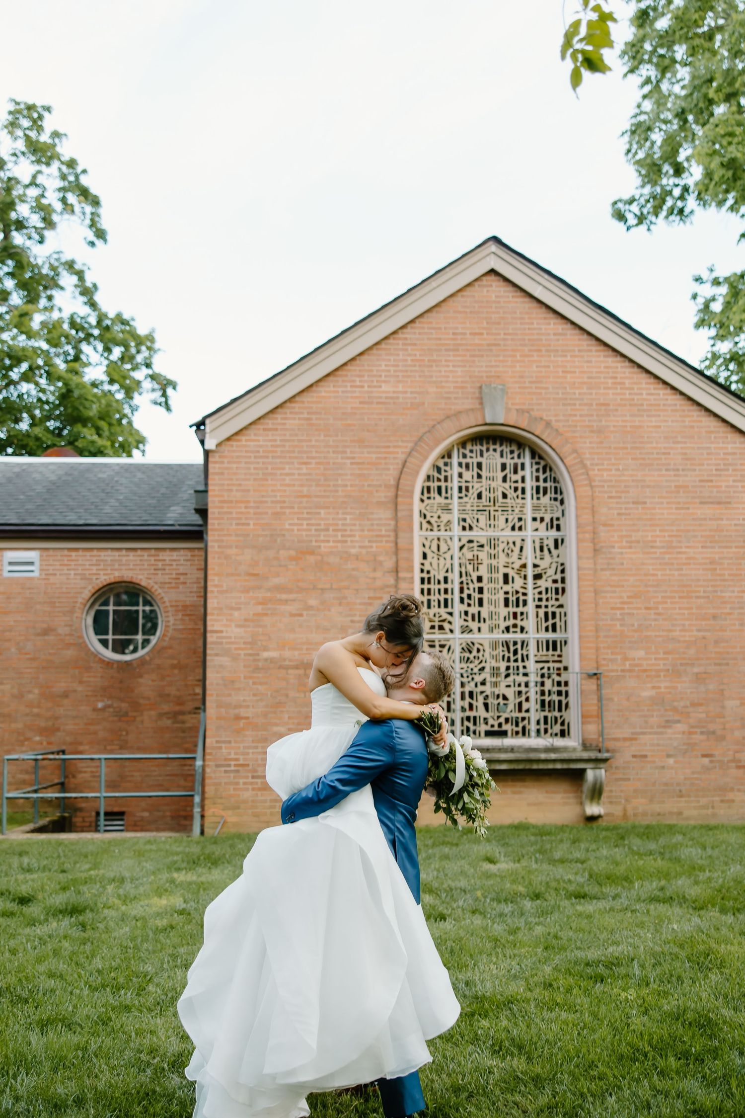 Wedding couple embracing outside brick building with decorative window.
