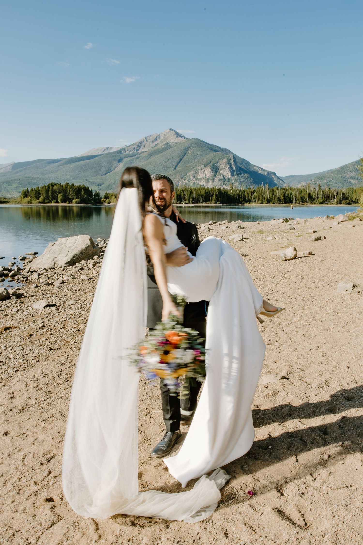 Wedding couple on rocky beach shore with mountain and lake backdrop.