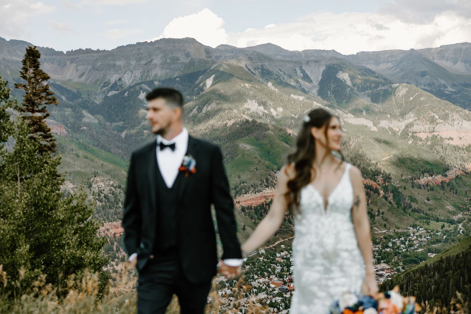 Newlyweds embrace on a mountain path with sweeping valley views and rugged peaks in the background.
