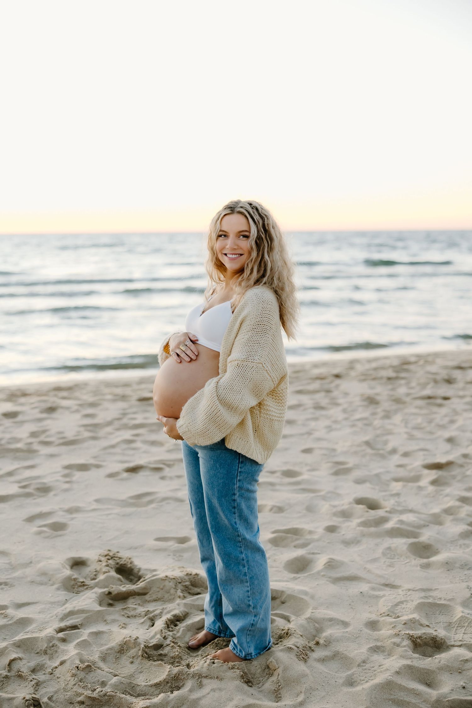 Maternity beach photoshoot with woman in cream sweater and jeans.