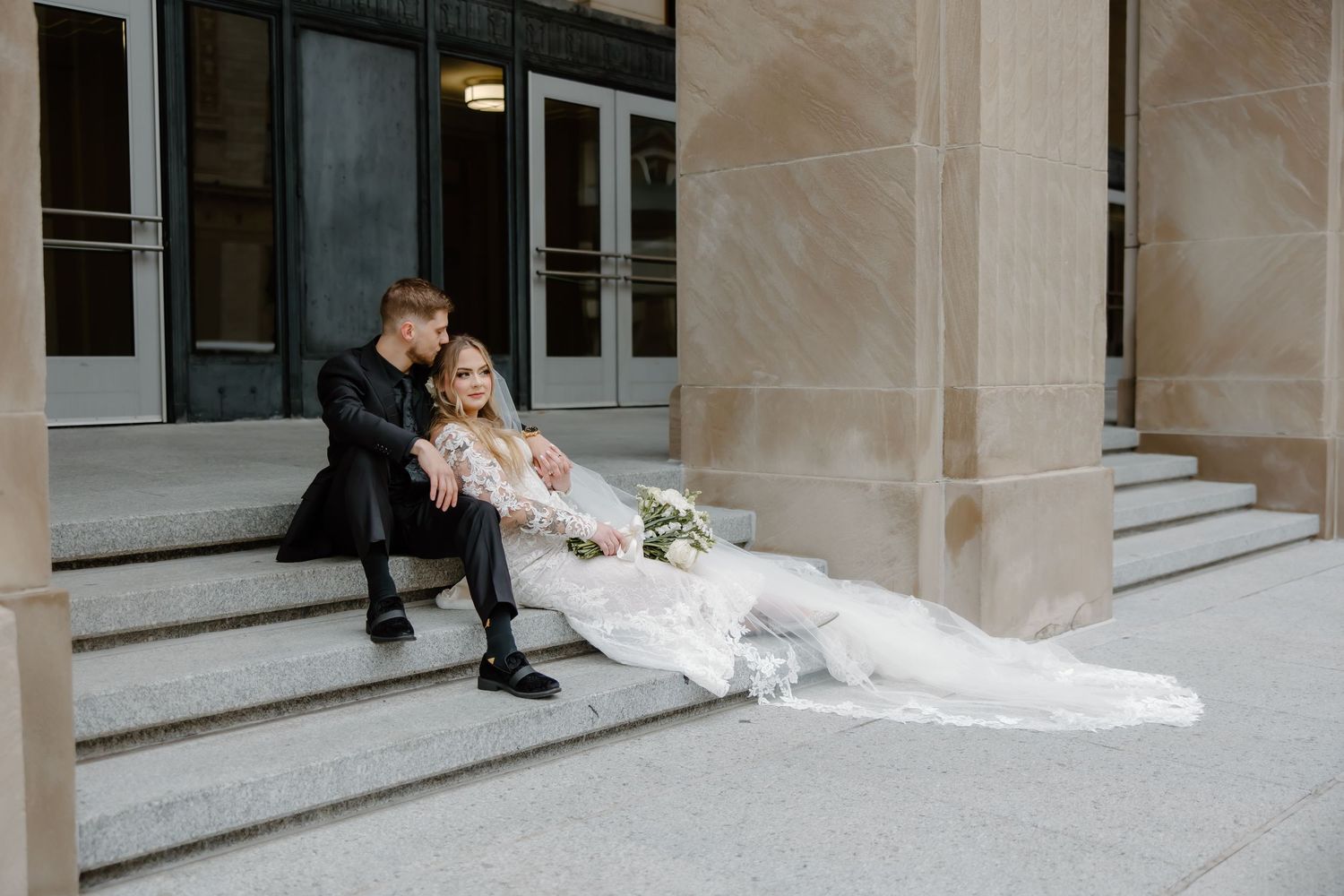 Bride in flowing white dress sits on stone steps beside columns.