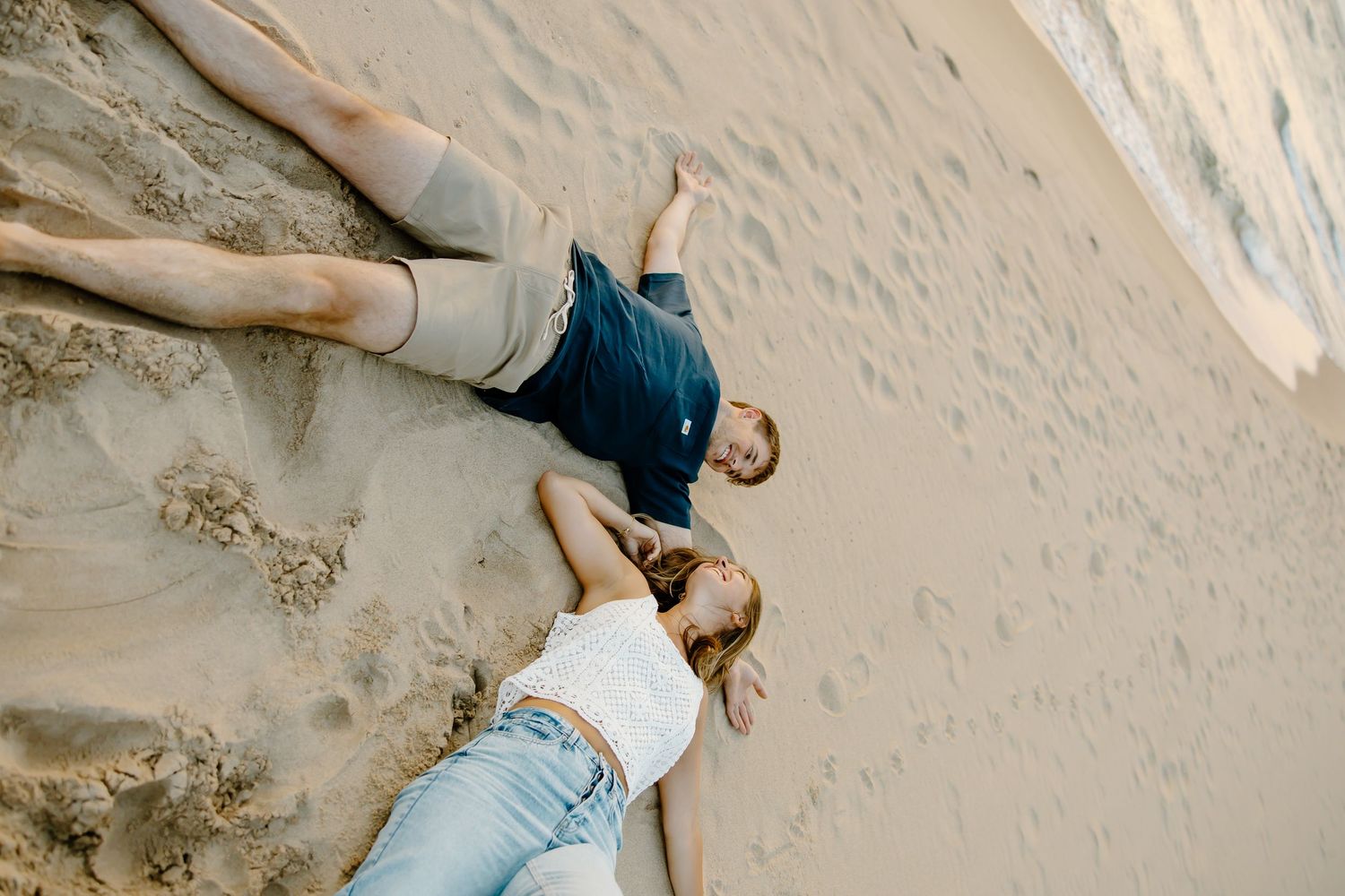 Two people lying on a sandy beach slope in casual attire.