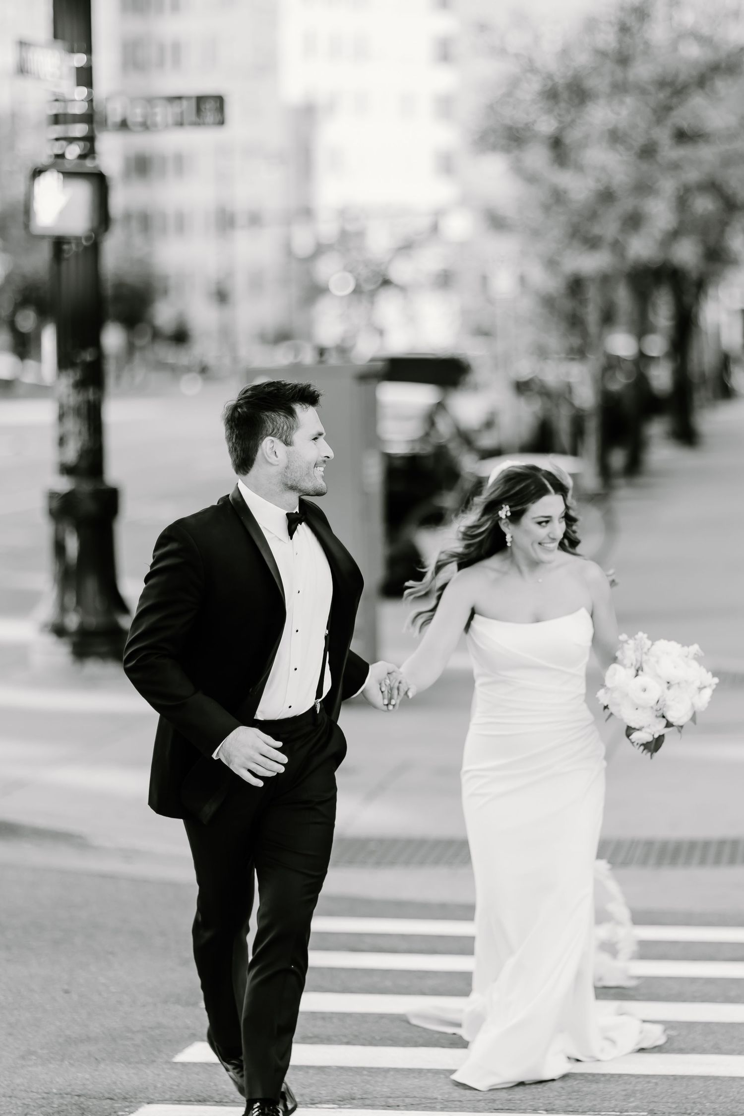 Black and white street photography of newlyweds walking playfully through city crosswalk.
