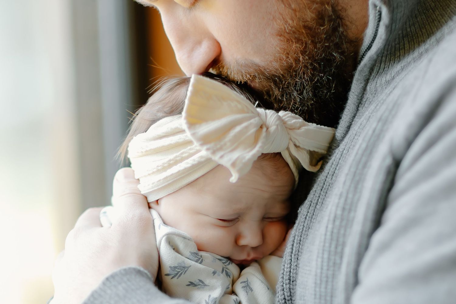 Parent in grey sweater cradling sleeping newborn against window light.