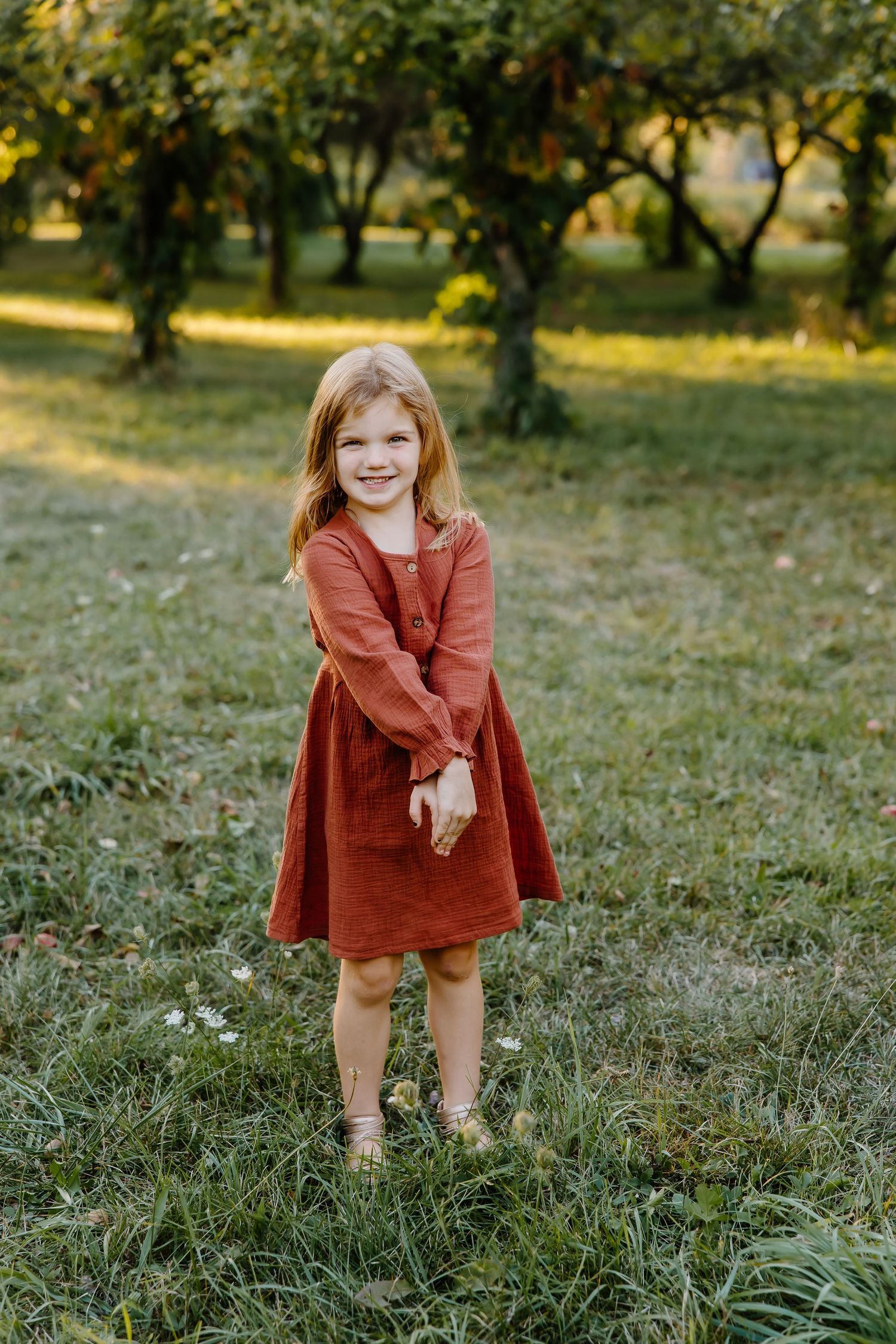 Young child in rust-colored dress stands sweetly in an orchard during golden hour.
