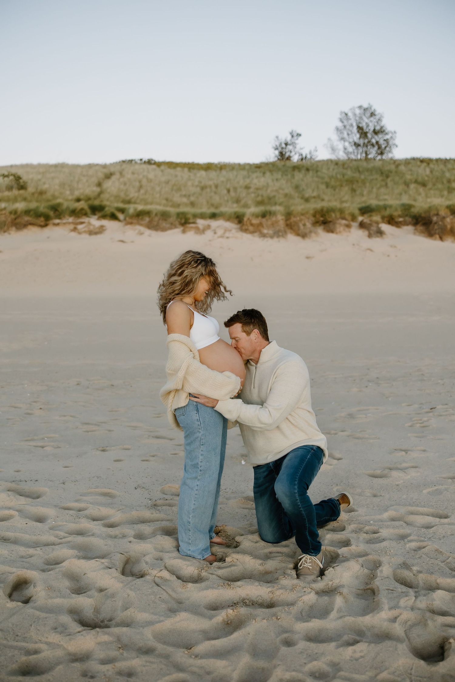 Romantic beach maternity photoshoot with couple in casual attire on sandy shore.