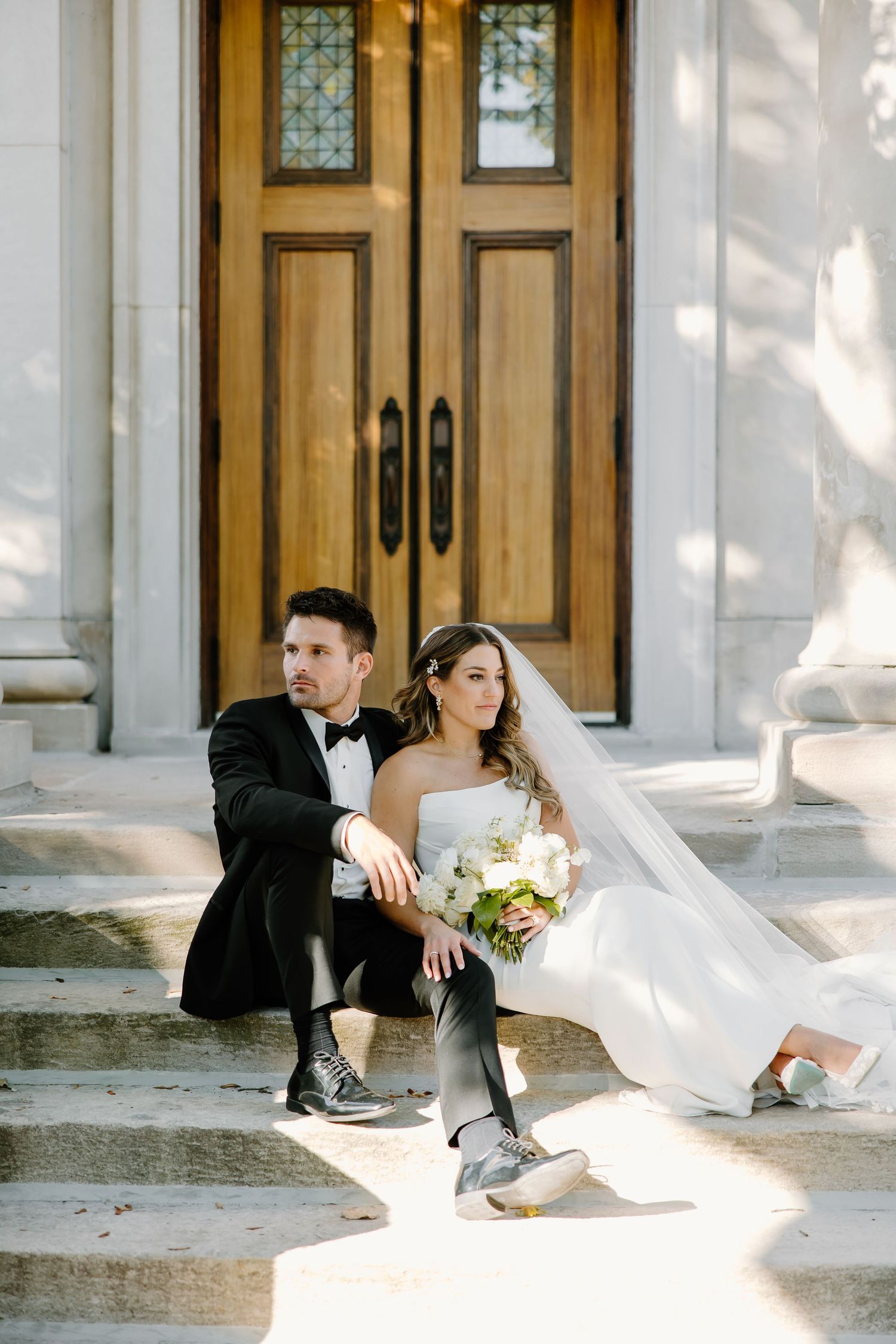 Wedding couple sitting casually on stone steps with sunlight filtering through columns.