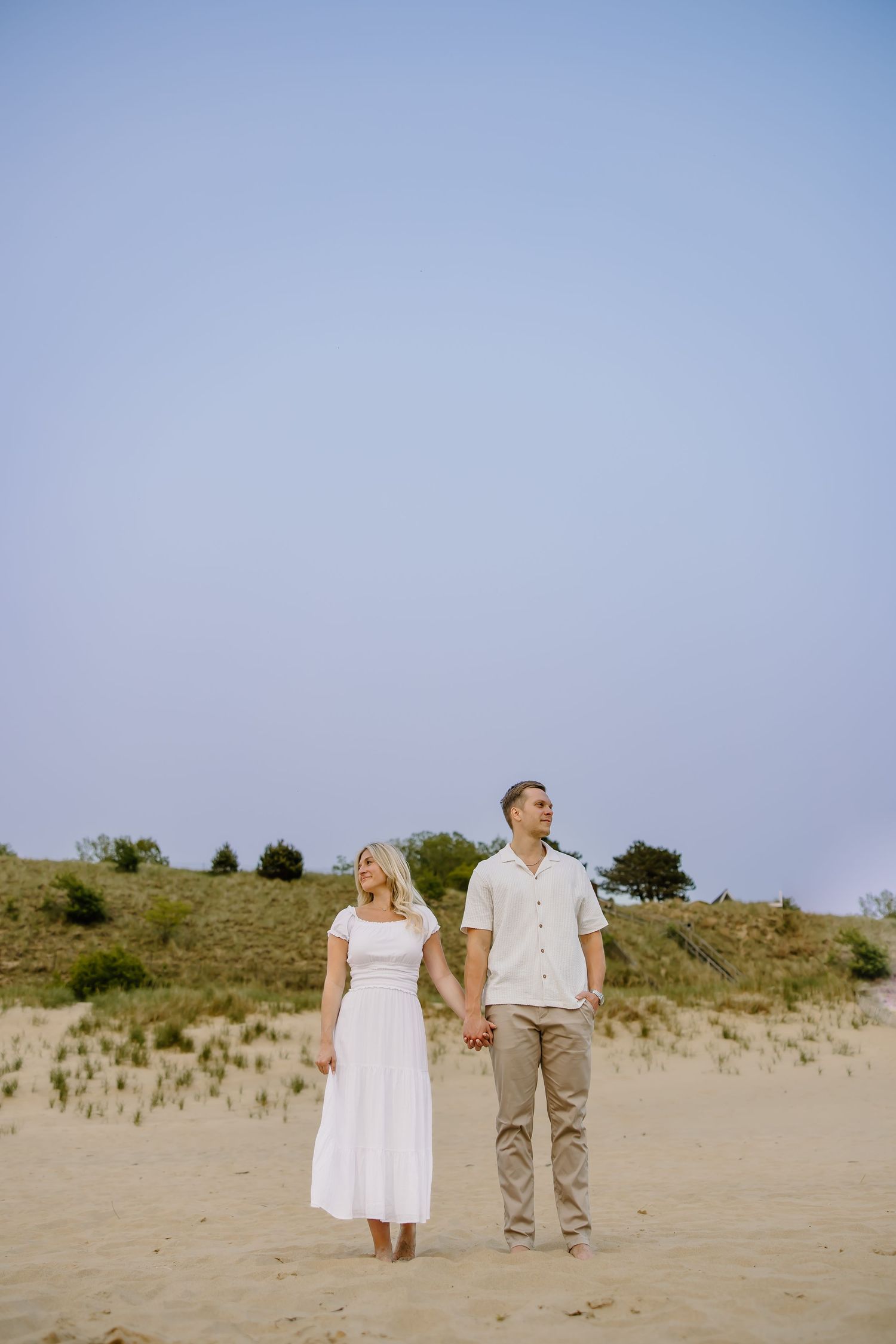 Couple in white and khaki standing together on sandy beach with dunes in background.