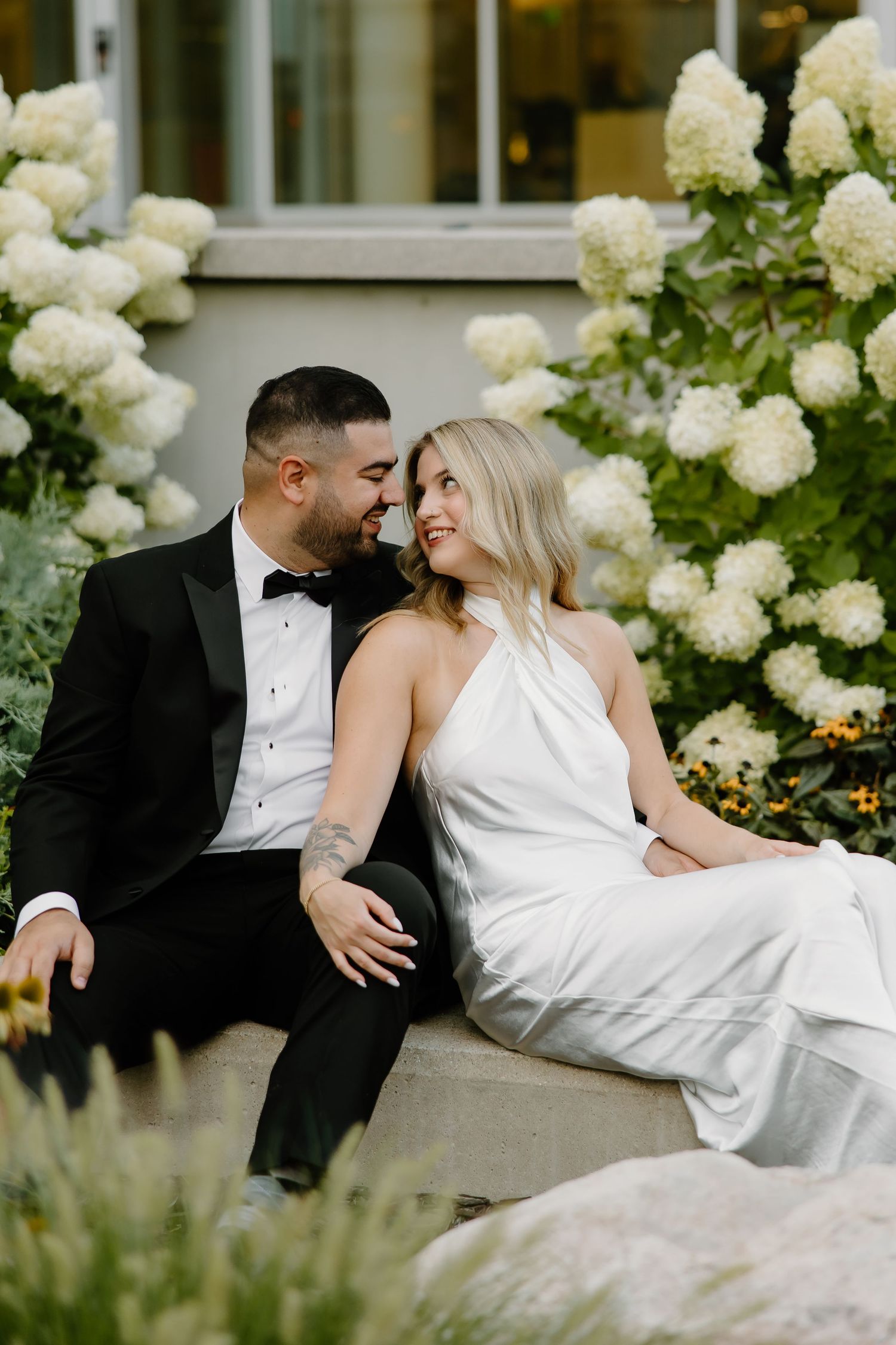 Couple lounges intimately by white hydrangea garden in formal attire.