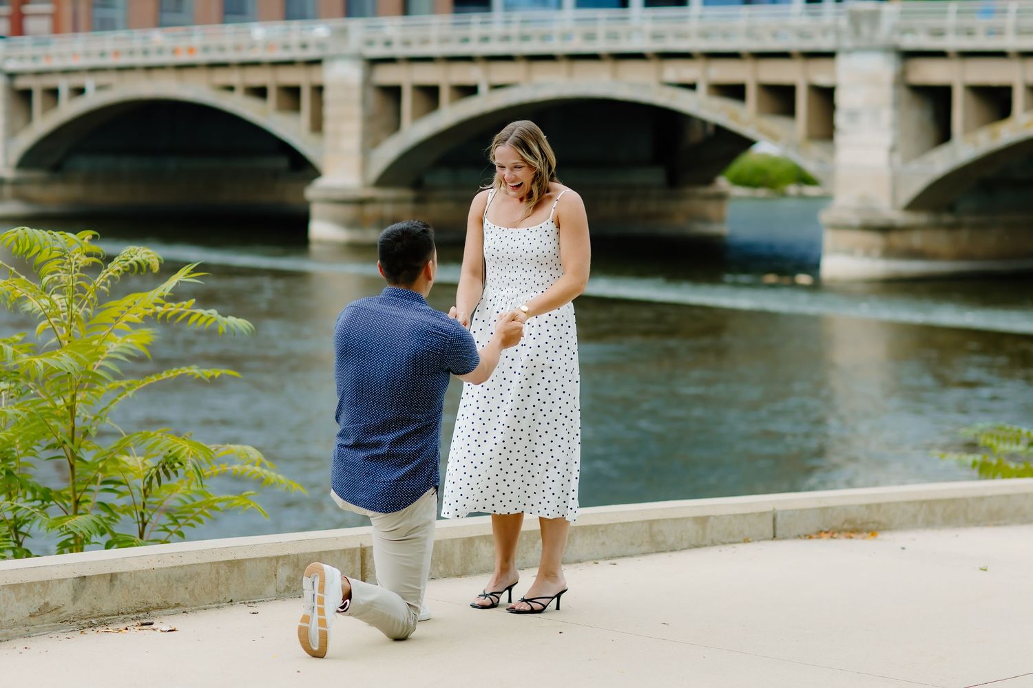 Surprise proposal moment by urban riverside with bridge in background.