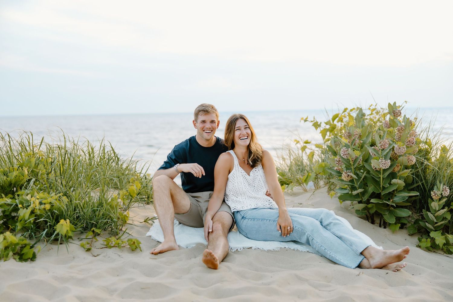 Romantic beach scene with couple lounging on white sand near flowering coastal plants.